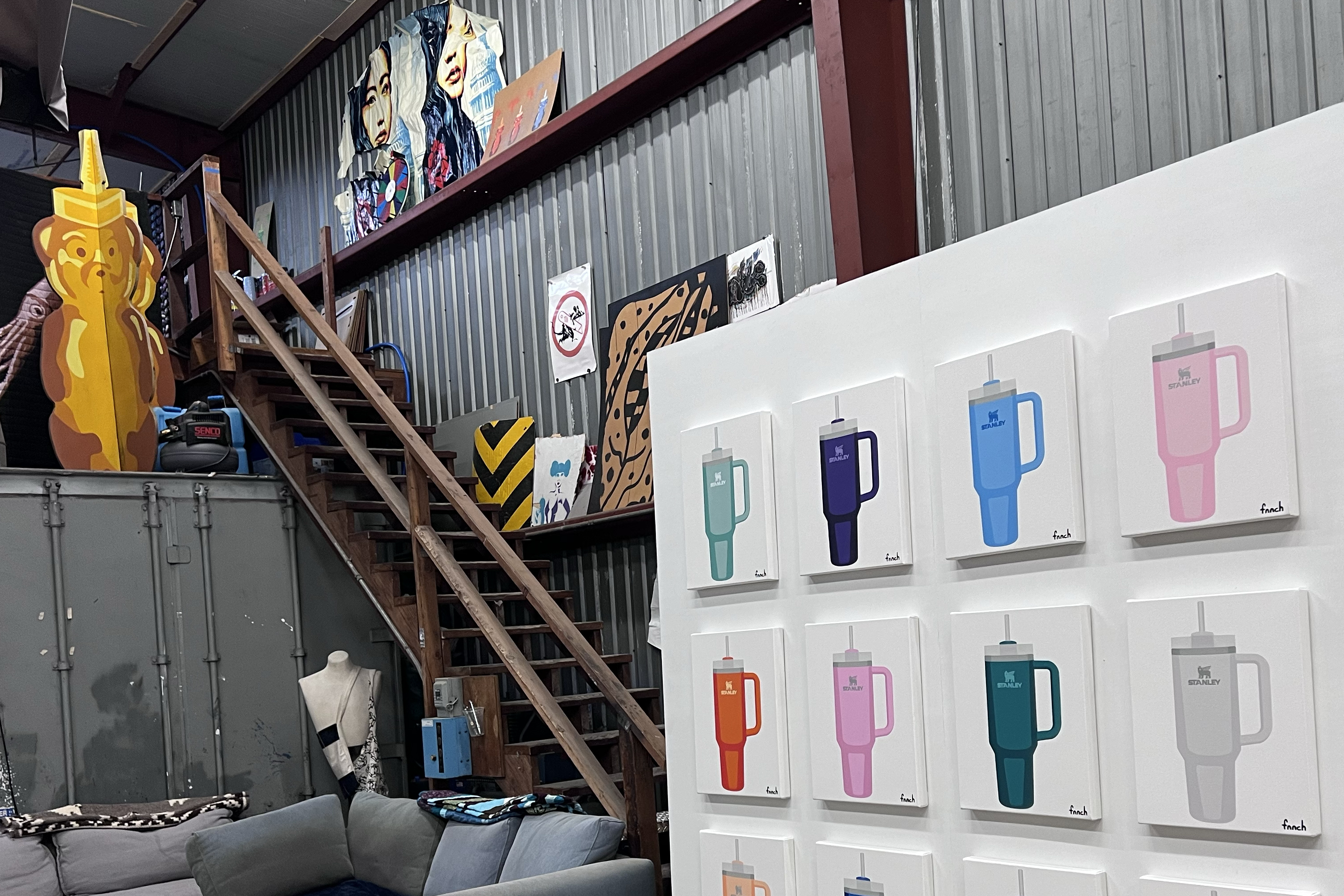 An eclectic room with artwork, colorful kettle illustrations on white canvases, a staircase, and a couch, all under a corrugated metal ceiling.