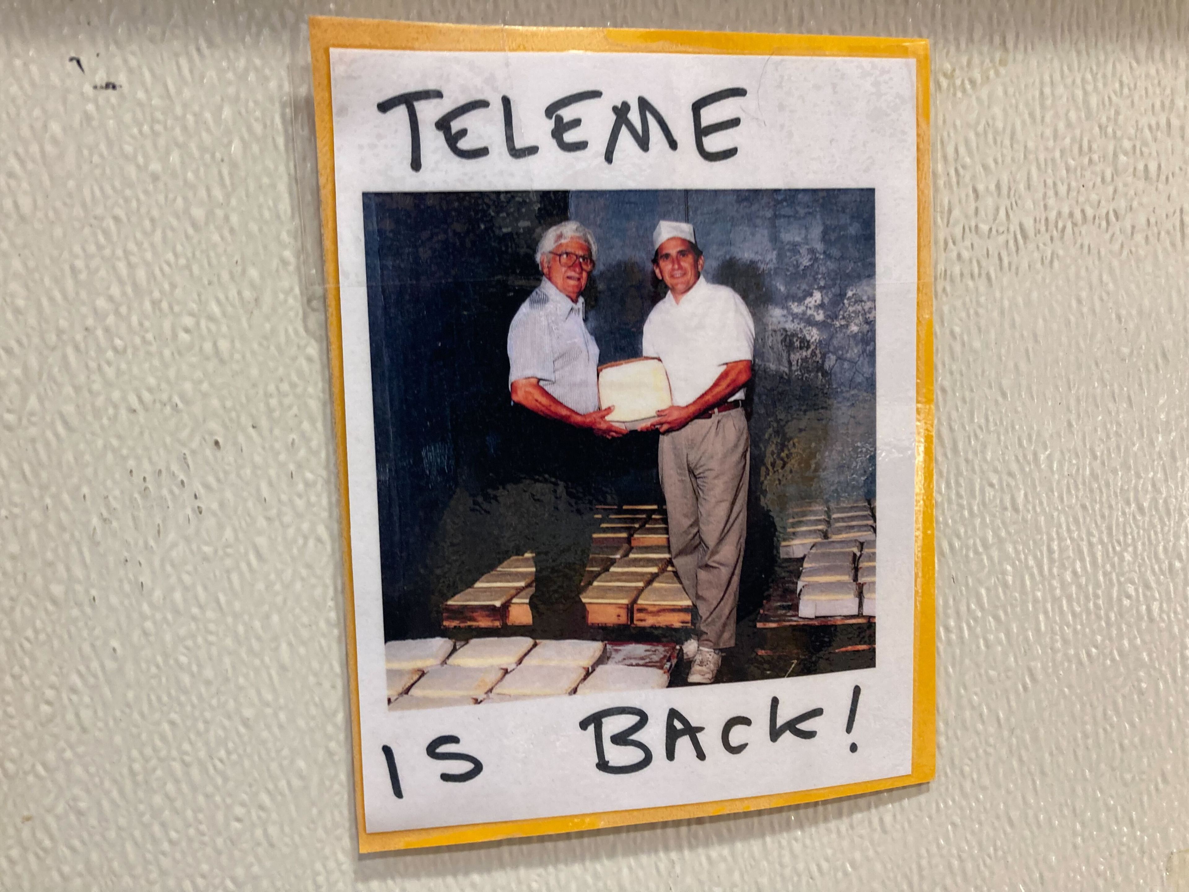 A sign reading &quot;teleme is back!&quot; is pinned to a wall and shows two cheesemakers holding a wheel of cheese.