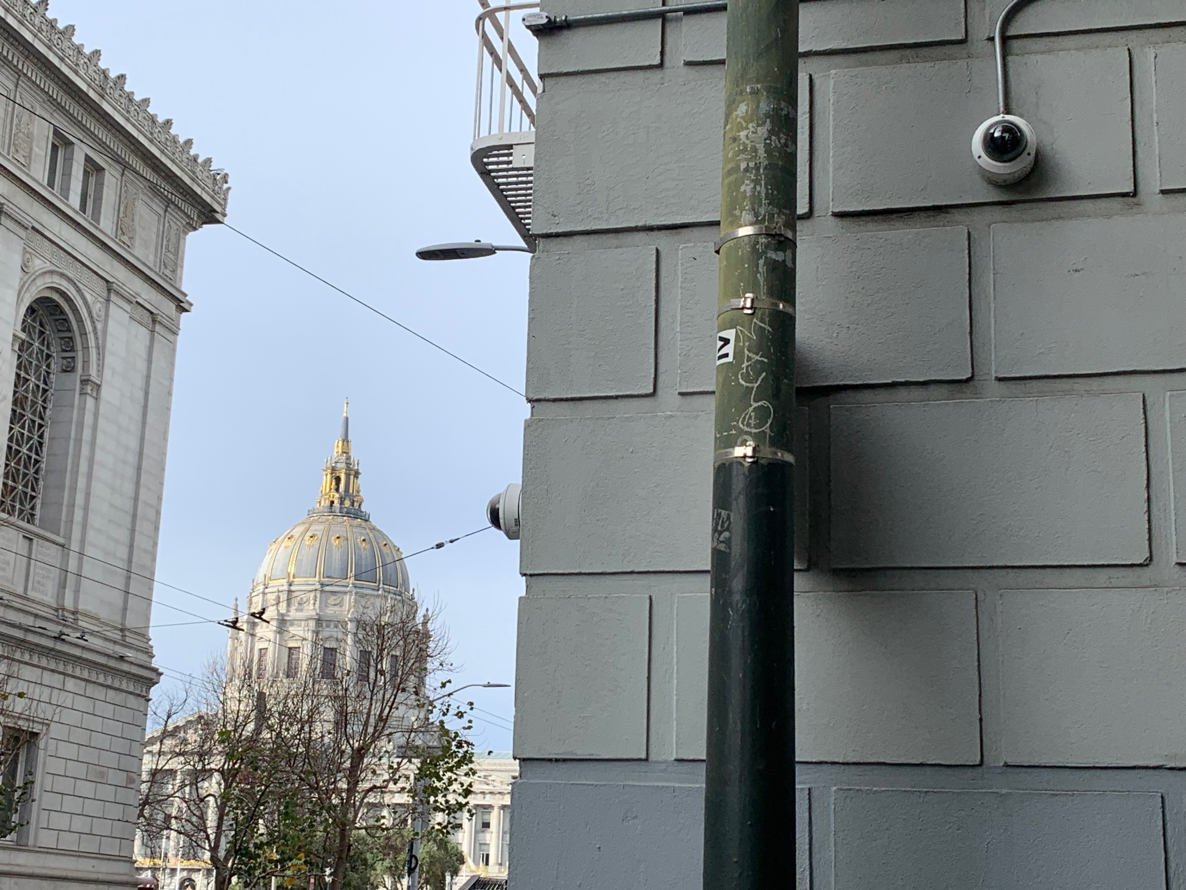 The image shows the dome of a grand building in the distance, partially obscured by trees. In the foreground is a building wall with a pole and a security camera.