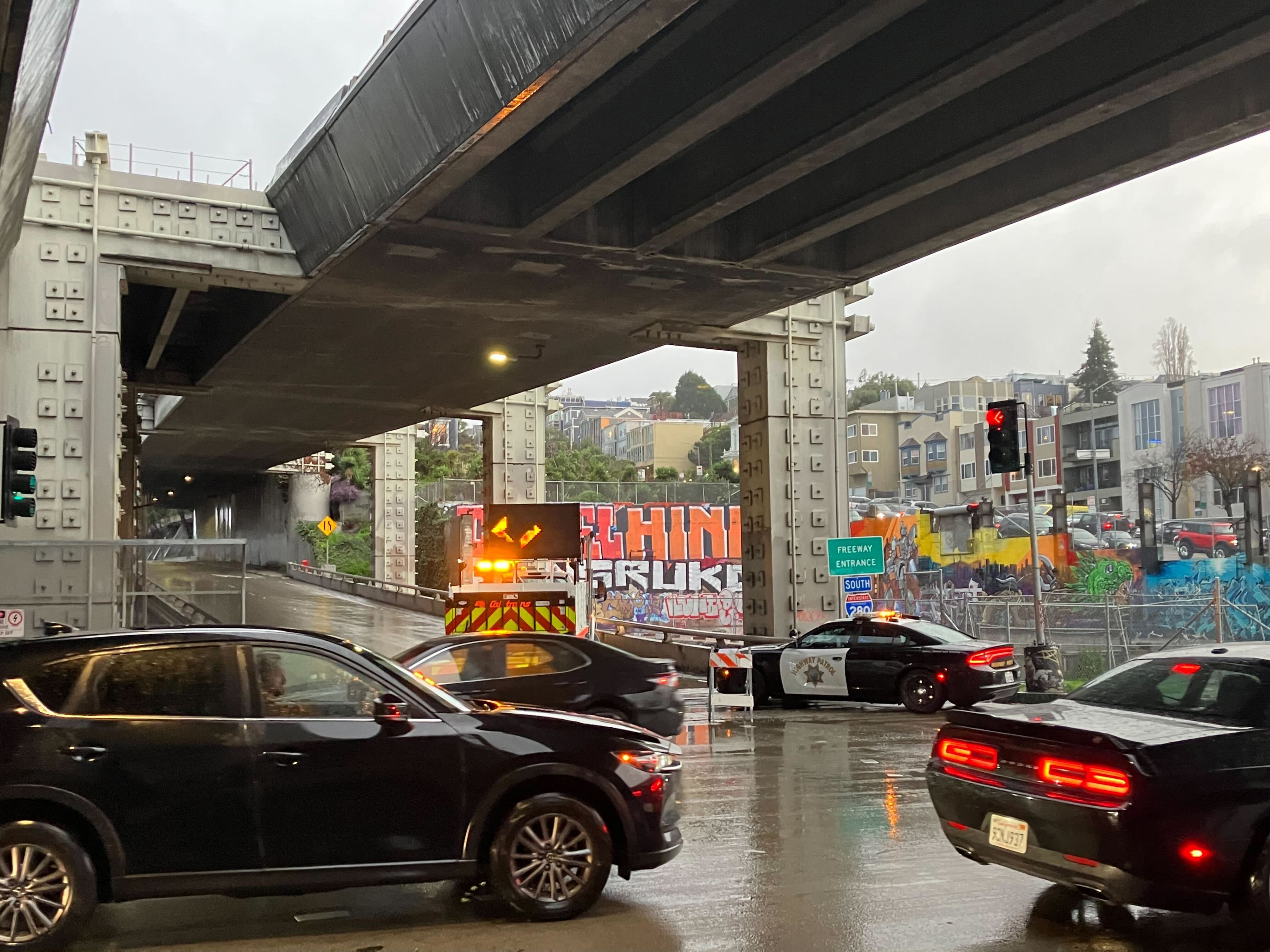 A rainy city scene under an overpass, with cars stopped at a traffic light and colorful graffiti on the sidewalls.