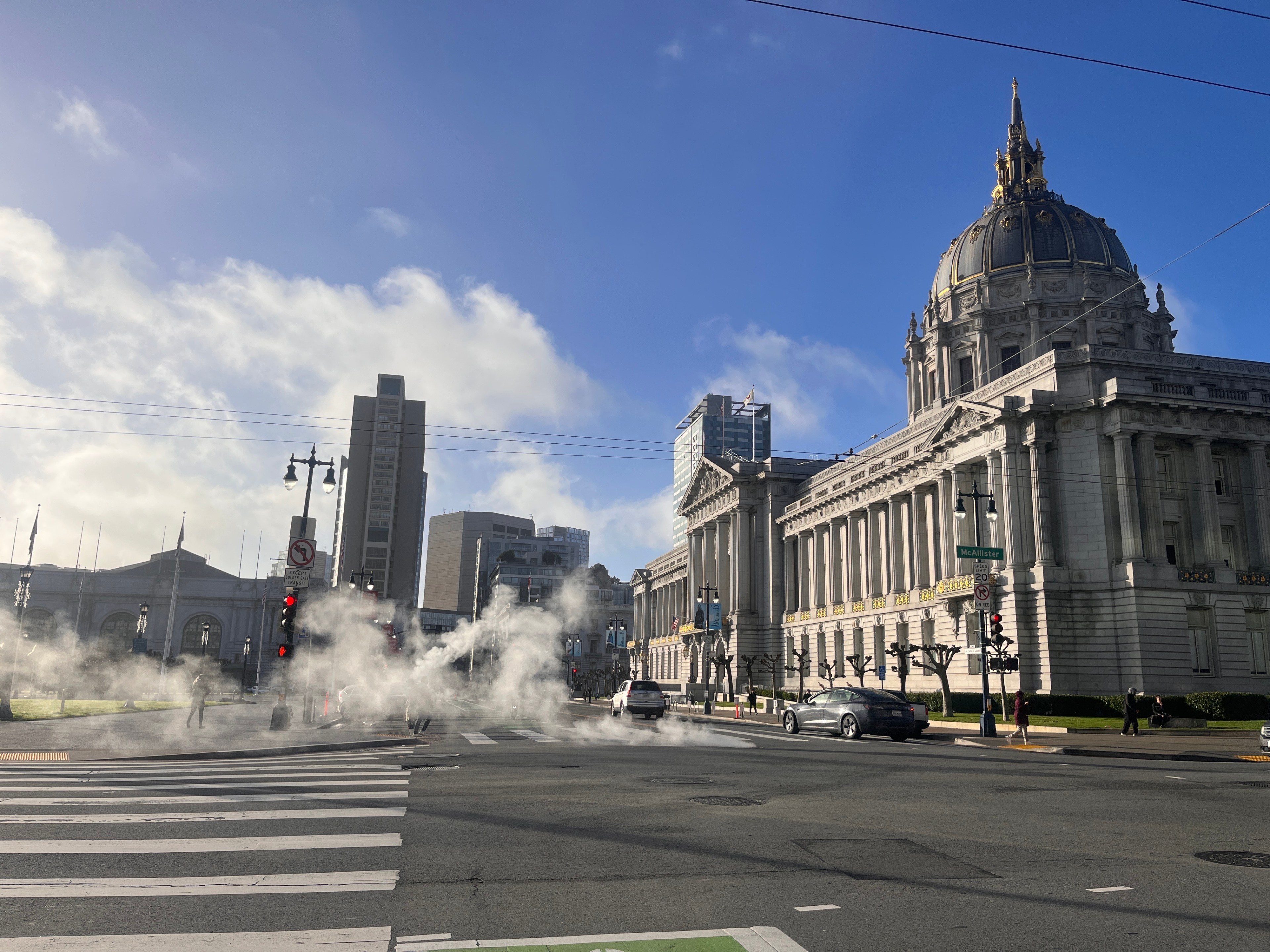 Steam rises from city street grates near a large domed building under a blue sky.