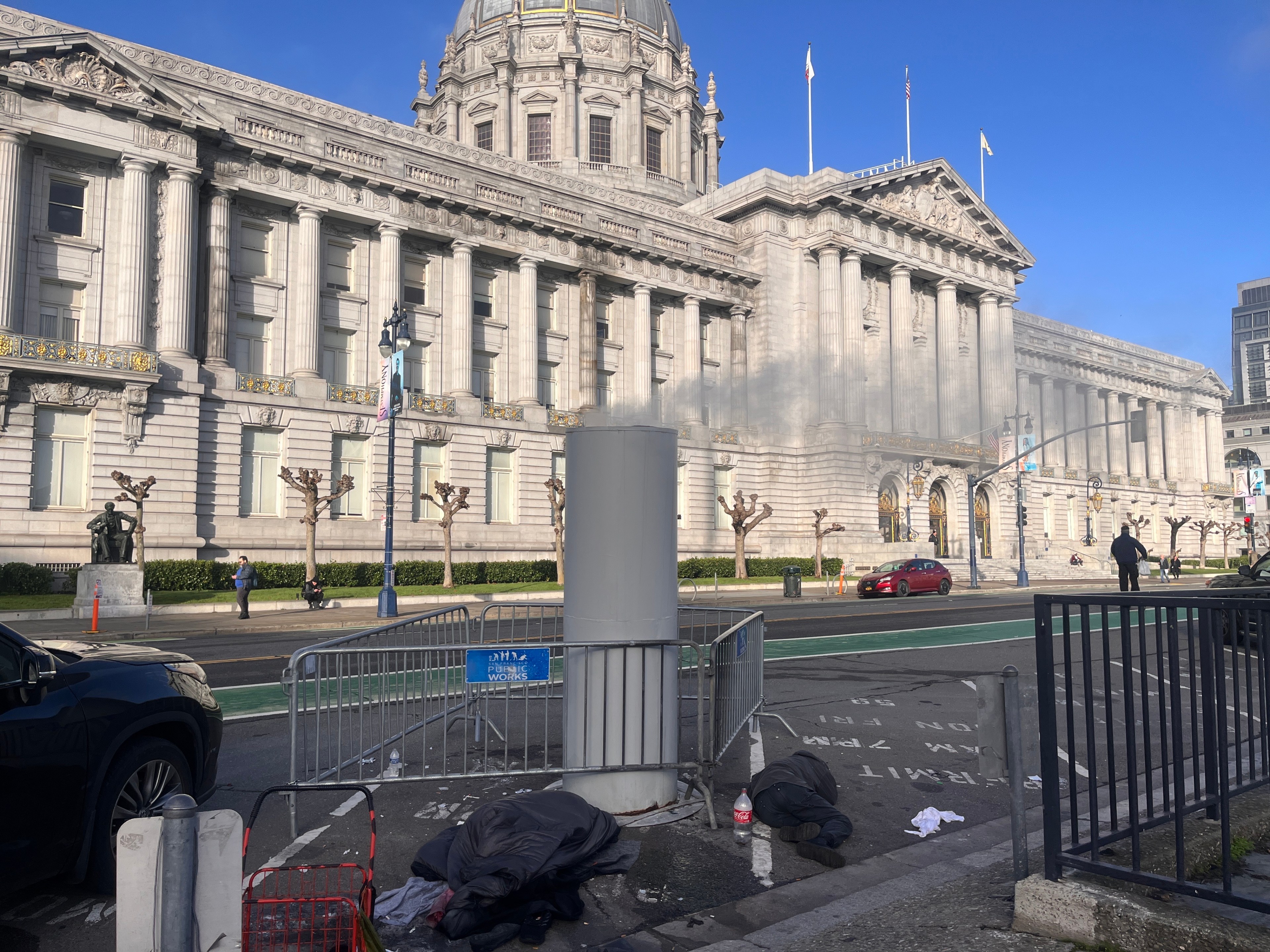 An imposing city hall with classical architecture and sculptures, with metal barricades and two people lying on the sidewalk covered with blankets.