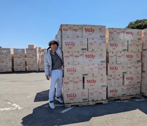 A man wearing glasses and a large smile and dressed in casual clothing stands on tiptoe besides several large wooden pallets containing cardboard boxes full of Pocky biscuit sticks.
