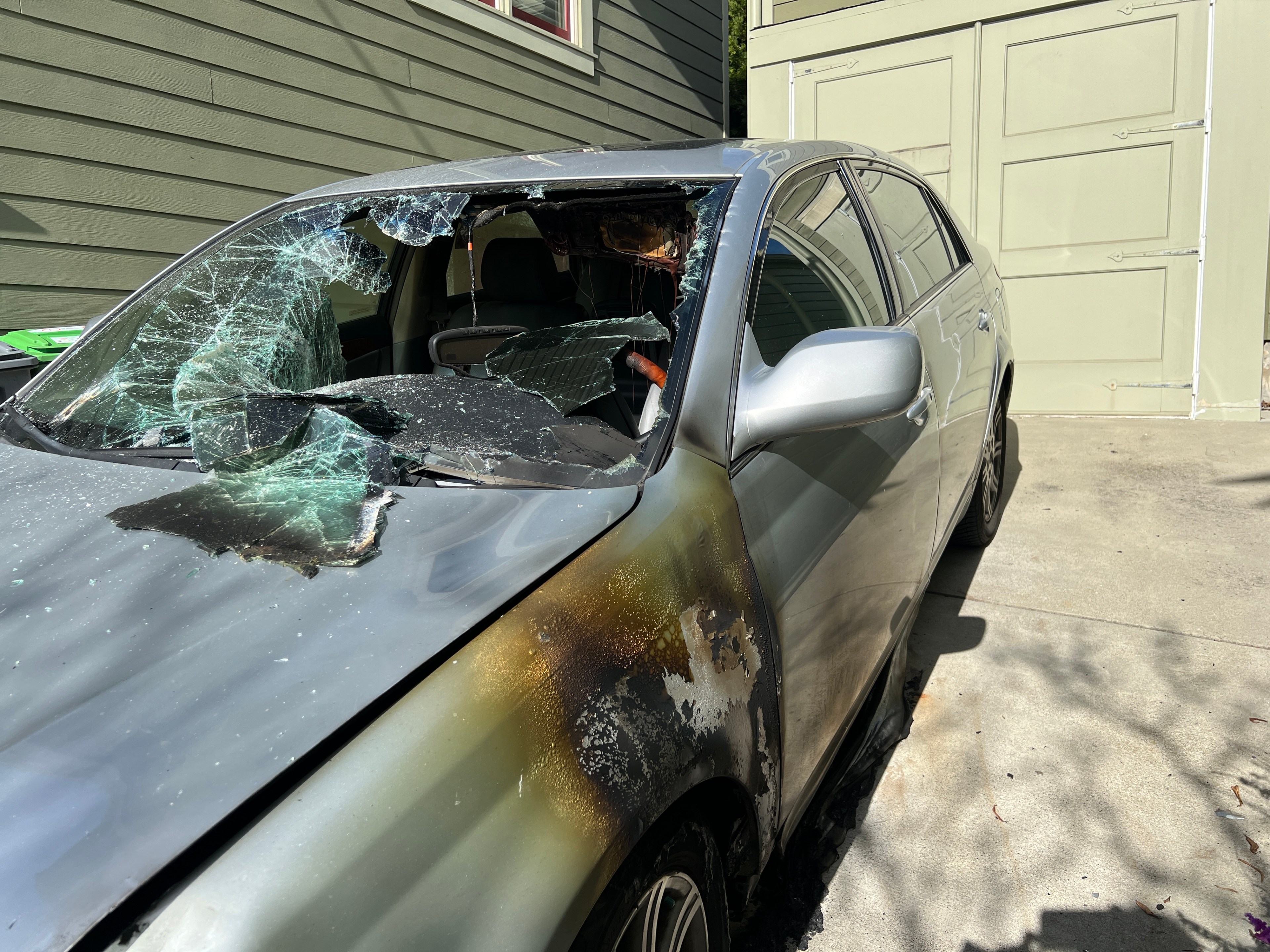 A gray car has a shattered windshield and a burned up side.