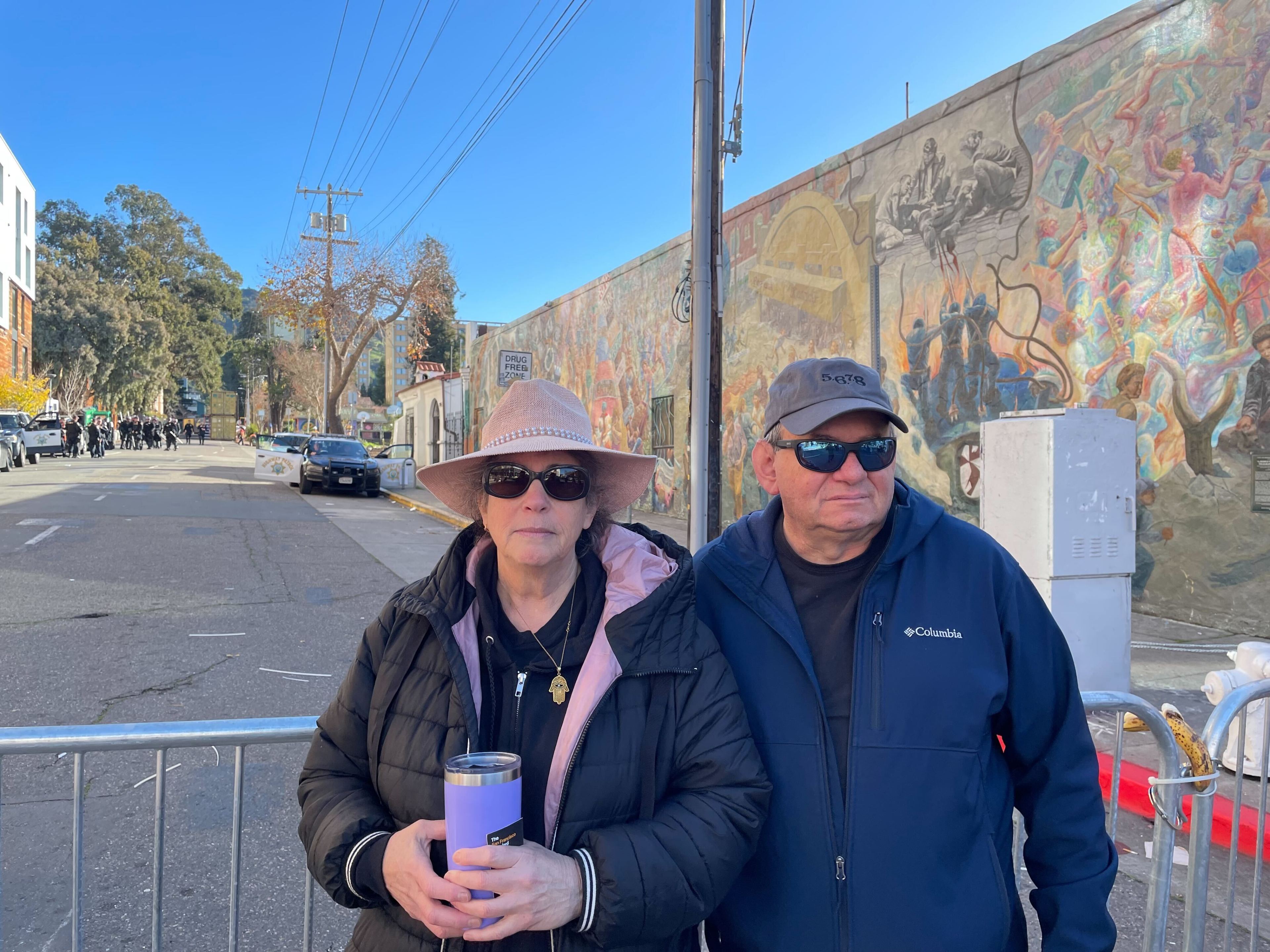 Two people wearing sunglasses, hats and warm dark-colored jackets stand beside a metal police barricade not far from a mural in the shady side of a street under sunny blue sky.
