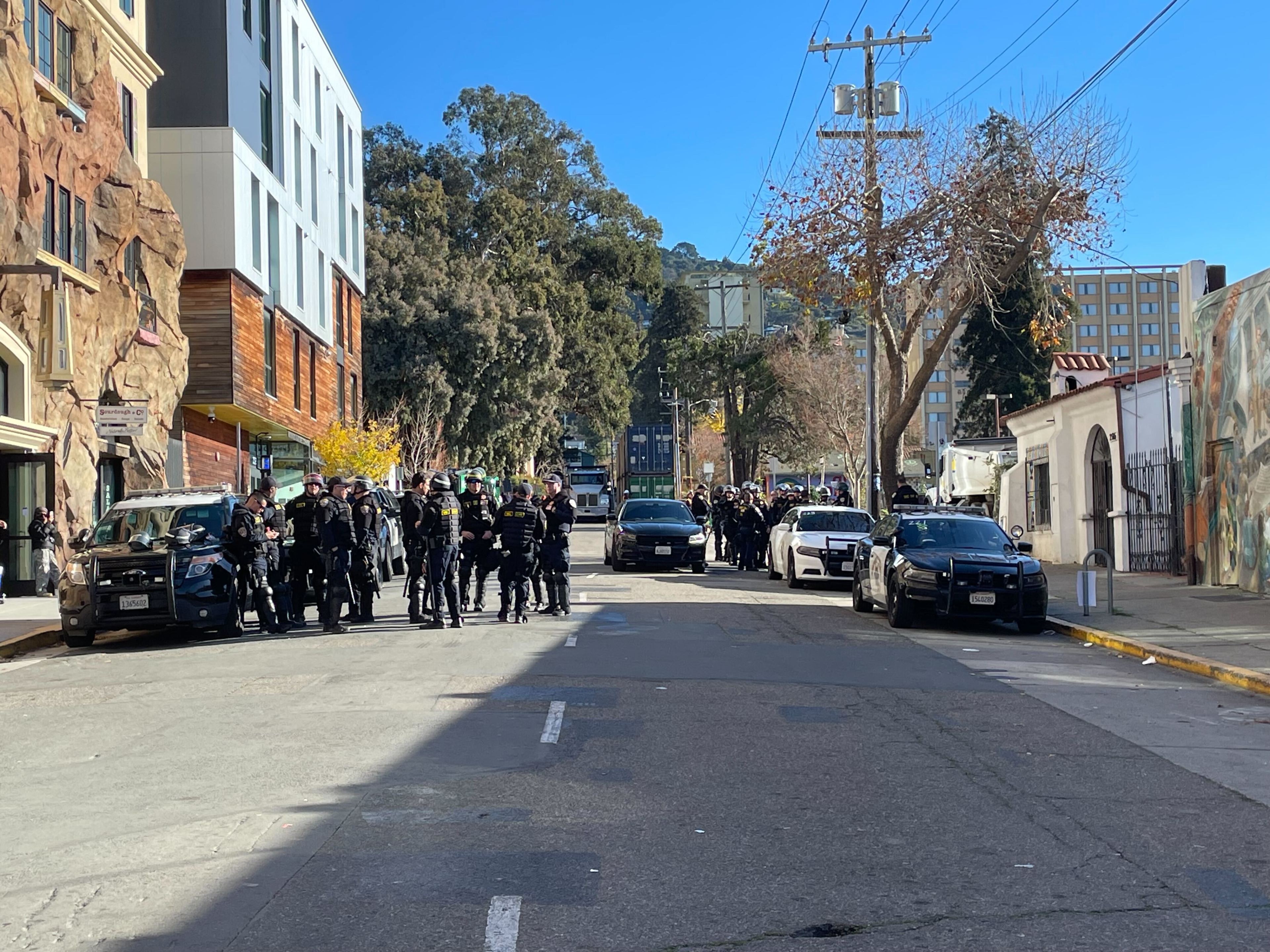 Multiple law enforcement personnel in tactical crowd-control gear gather near a parked patrol vehicle on the sunny side of a city street.