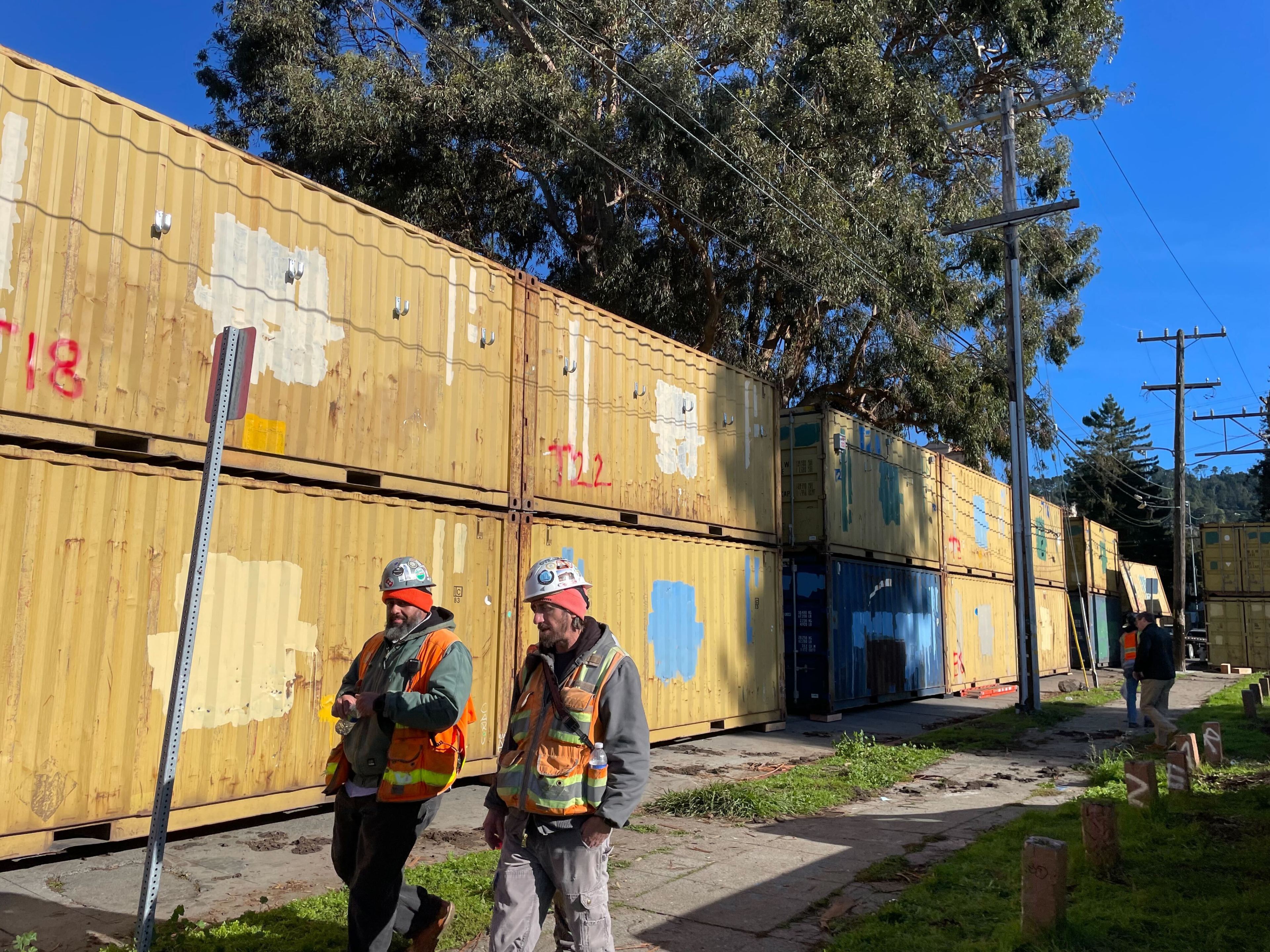 Two construction workers in high visibility vests and helmets walk and talk along a sidewalk beside a row of yellow double stacked shipping containers in a street.