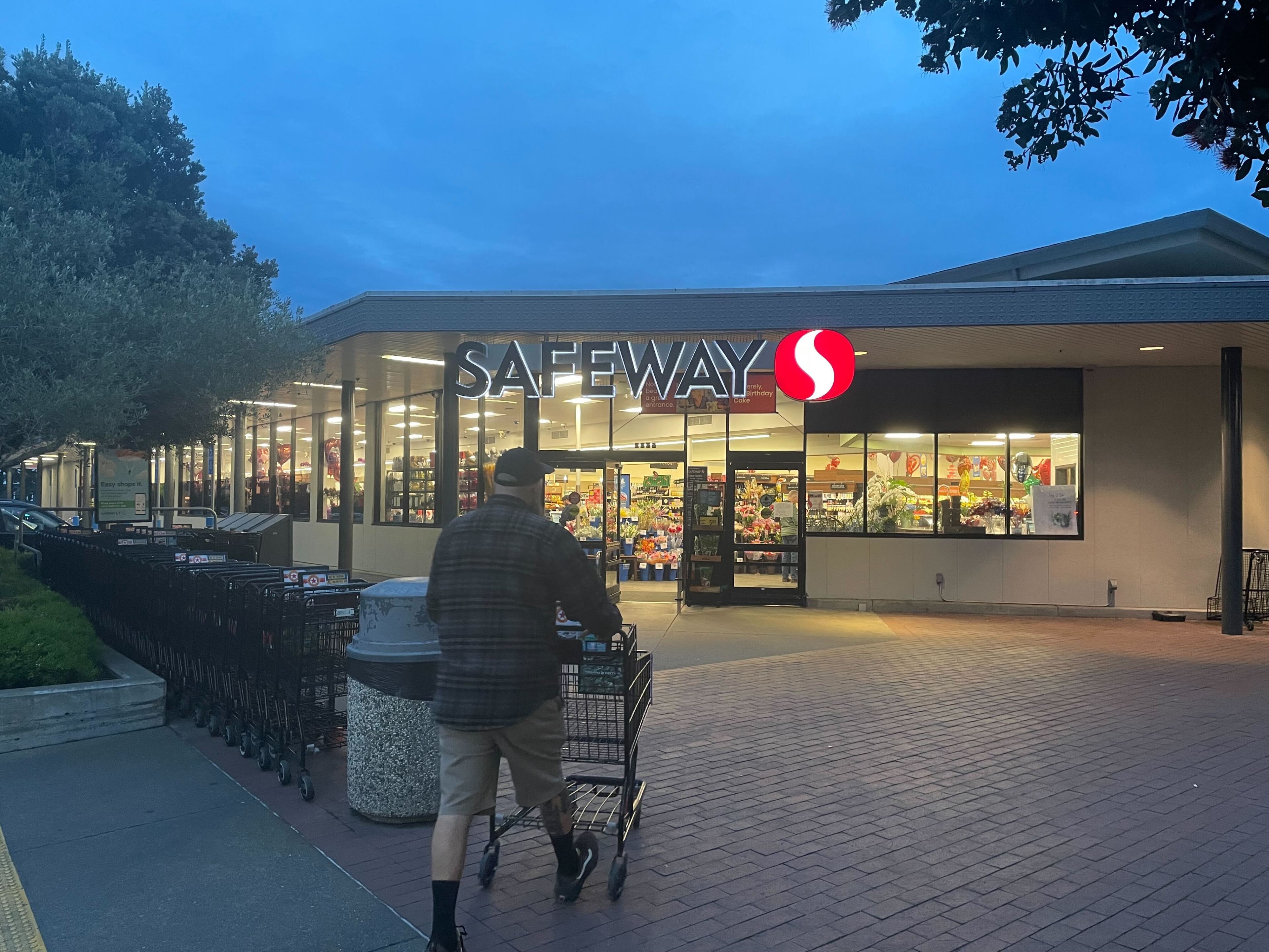 A person pushes a cart towards a Safeway grocery store at dusk, with shopping carts and bright store lights visible.