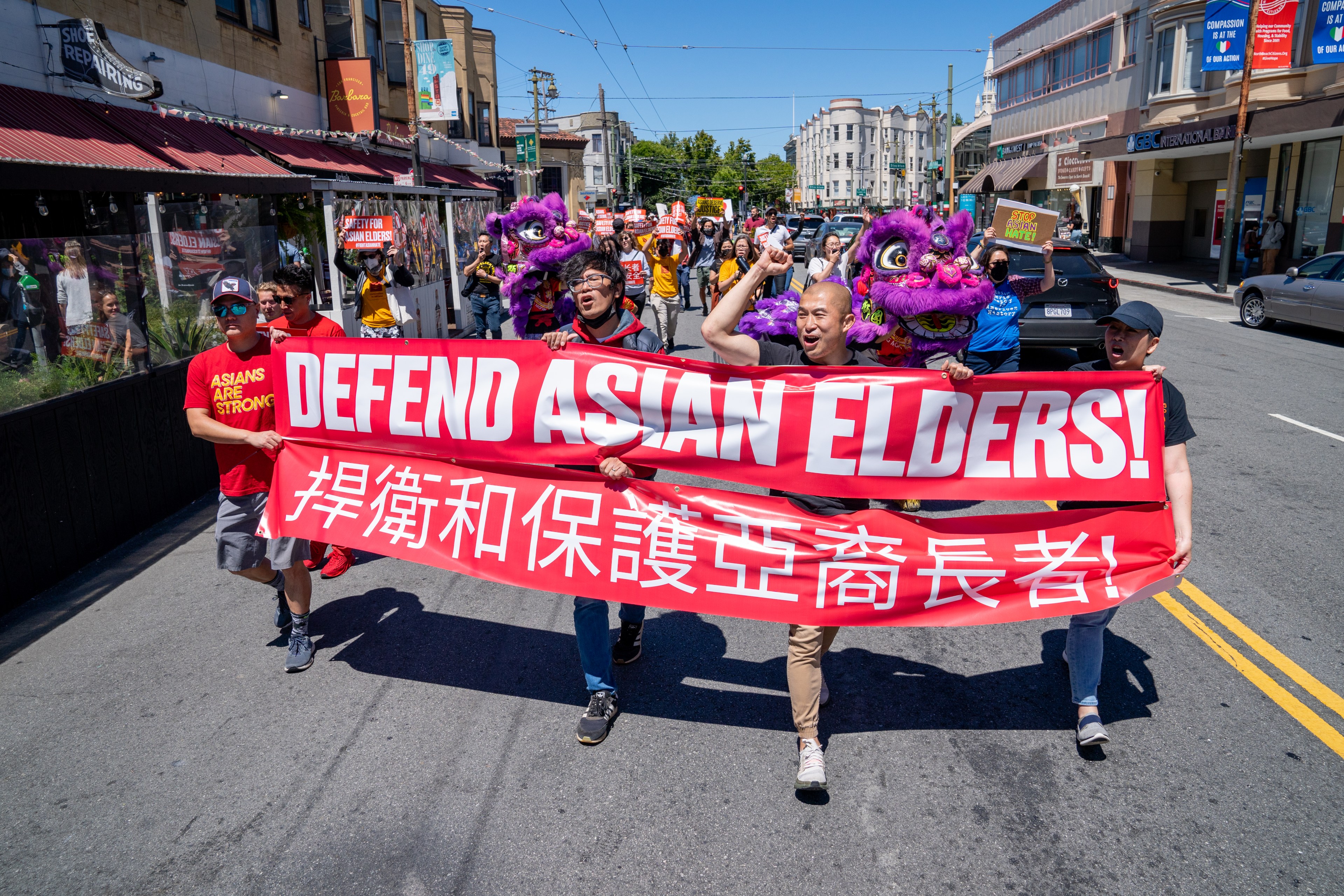 A group marches with a banner reading &quot;DEFEND ASIAN ELDERS!&quot; in both English and Chinese.