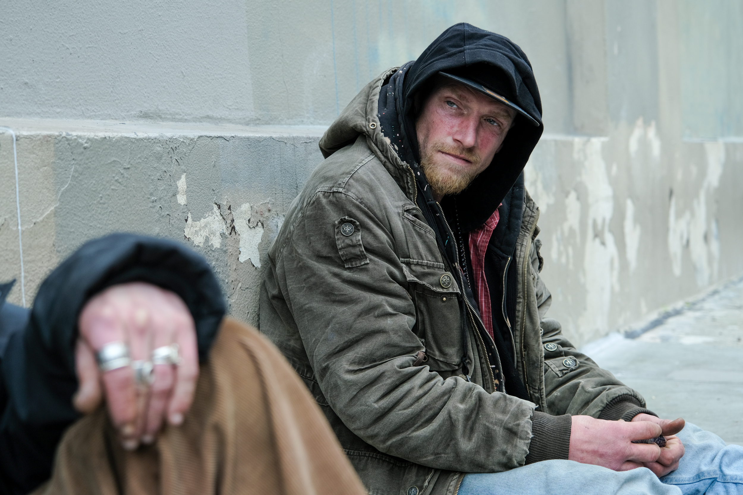 A man in a hooded jacket sits against a wall, looking at the camera, another person's hand in the foreground.
