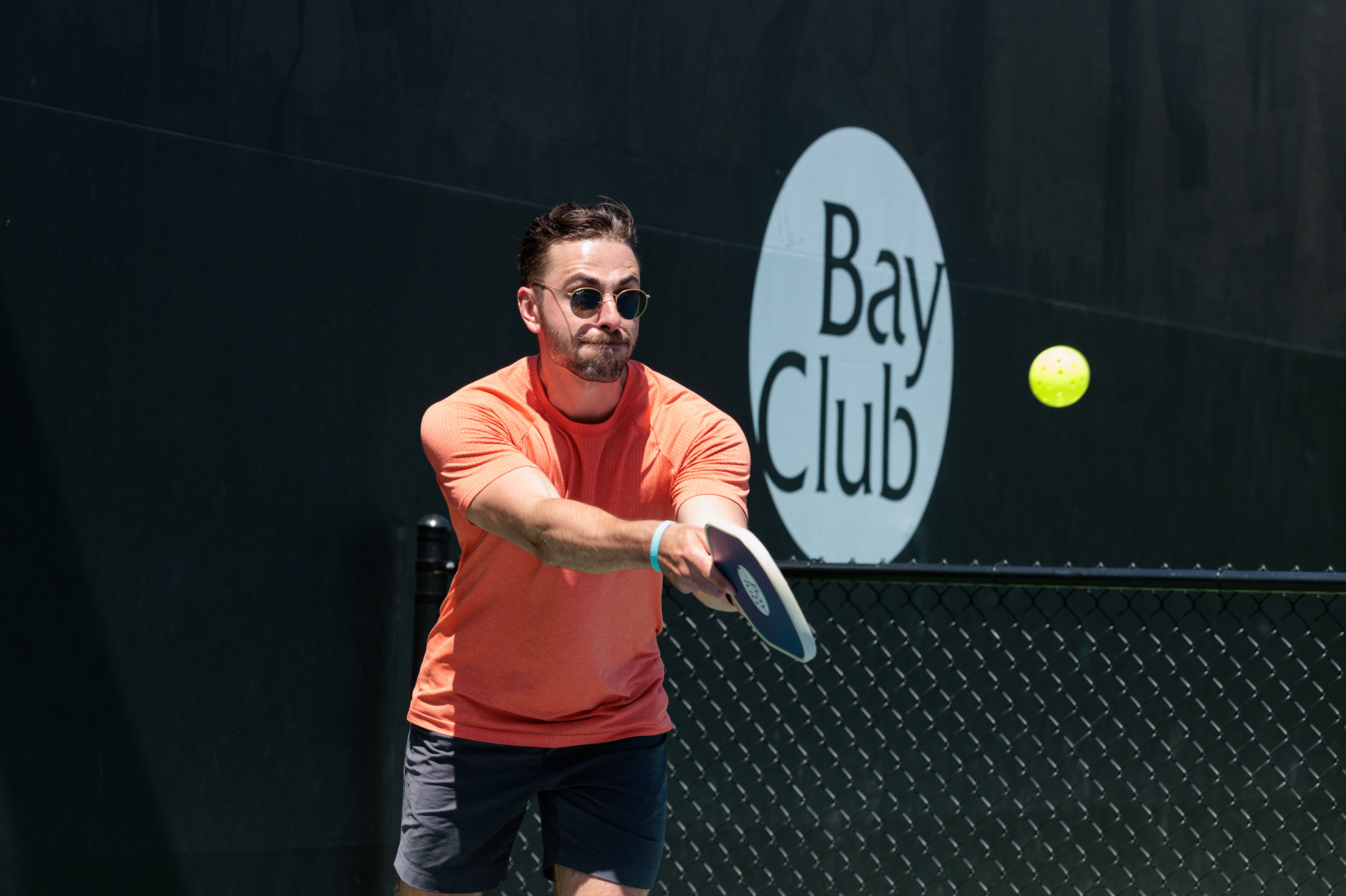 A man plays pickleball, in orange shirt and sunglasses, with a focused expression.