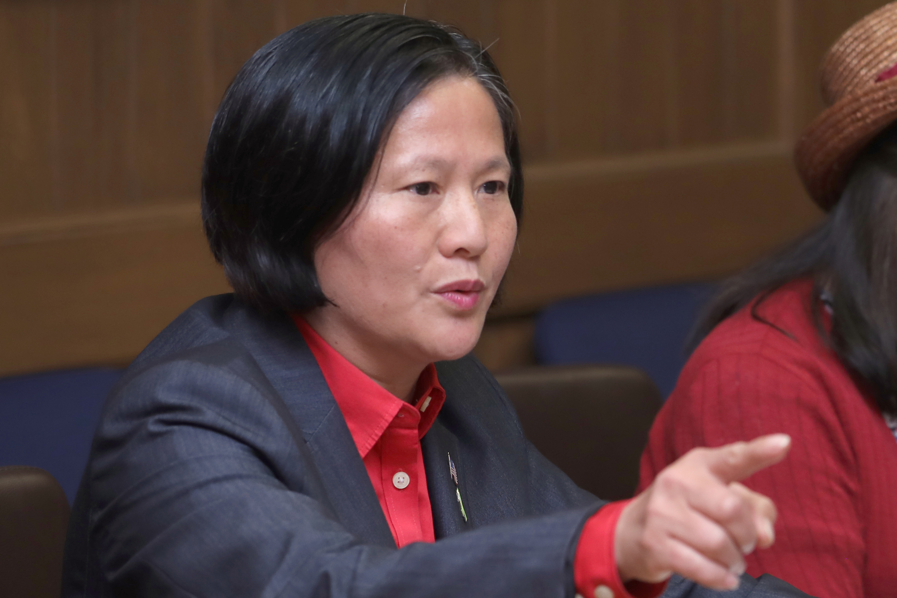 A woman with short black hair, wearing a red blouse and dark blazer, gestures while speaking to someone off-camera.