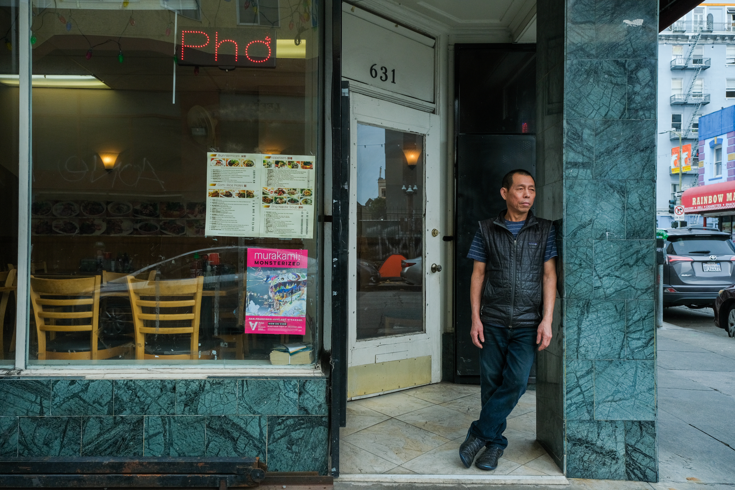 A man stands by an open door of a Pho restaurant, with a menu and a colorful sign in the window.