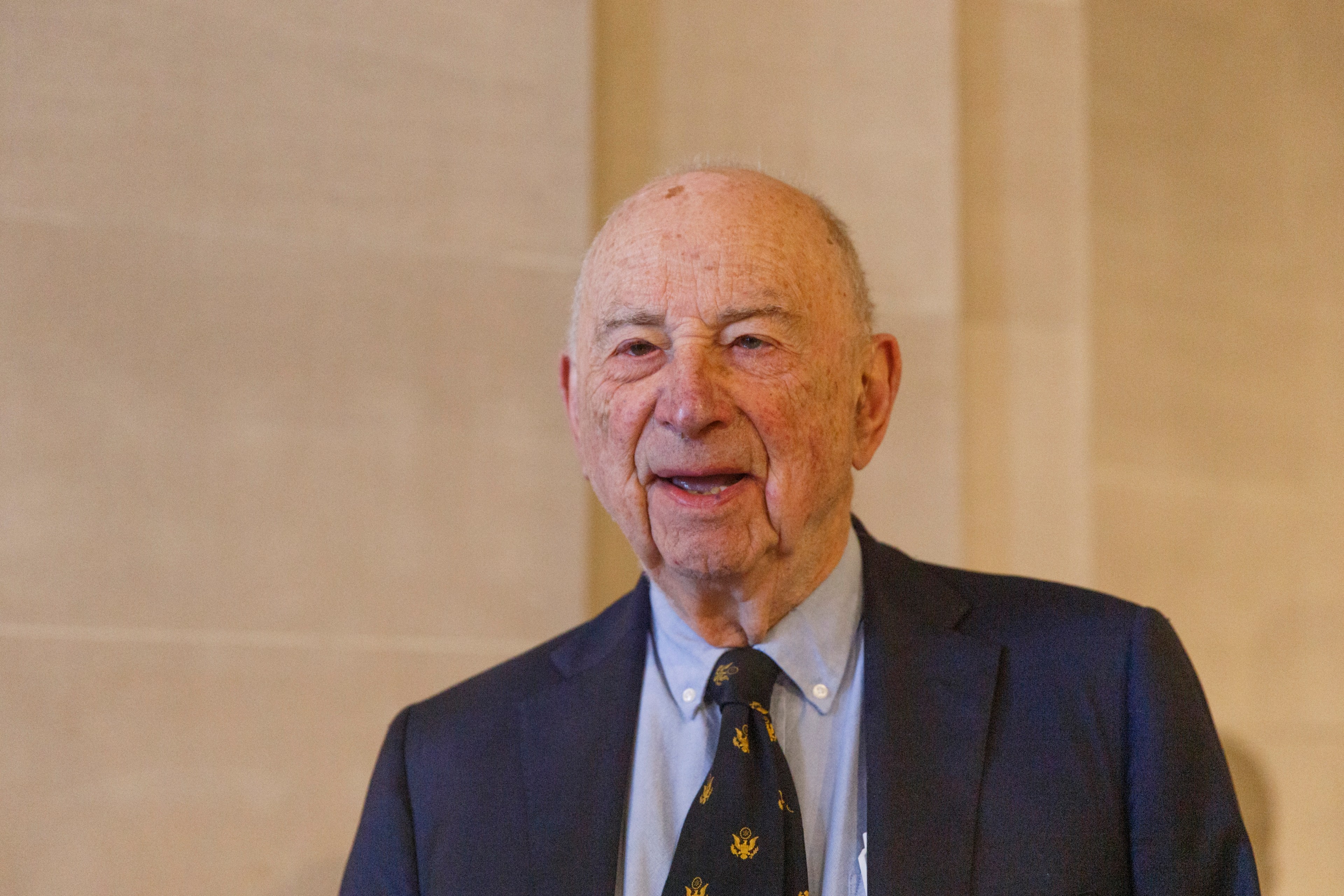 An elderly man with a warm smile, wearing a suit and tie, stands before a light background.