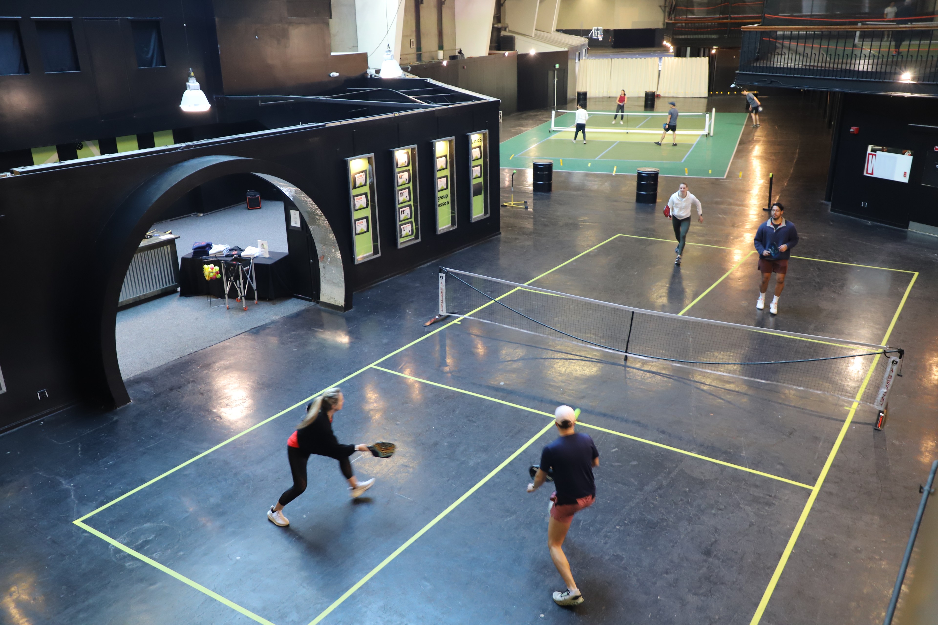 Four people play pickleball indoors with spectators above. Yellow lines mark the court on a dark floor.