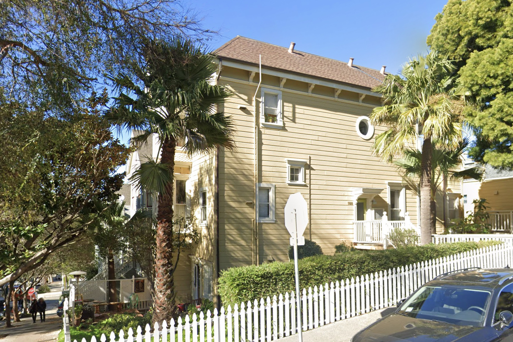 A beige two-story house with a white picket fence, surrounded by palm trees, under a clear blue sky.