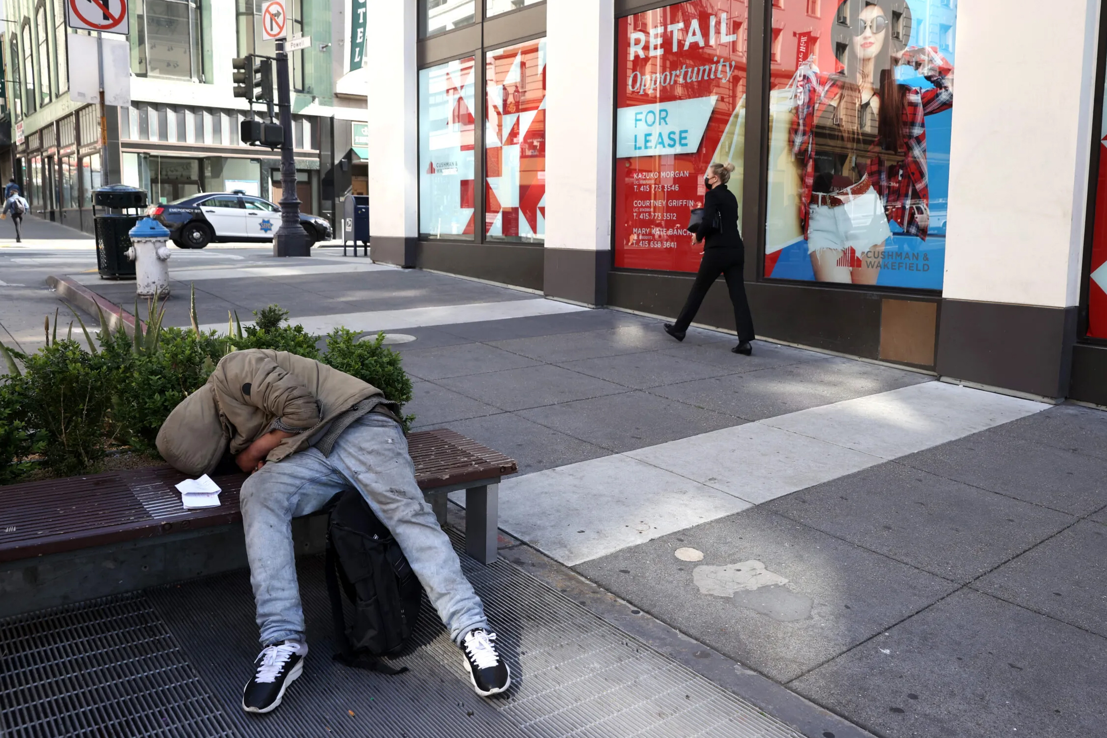 A homeless person sleeps on a bench in front of closed retail stores