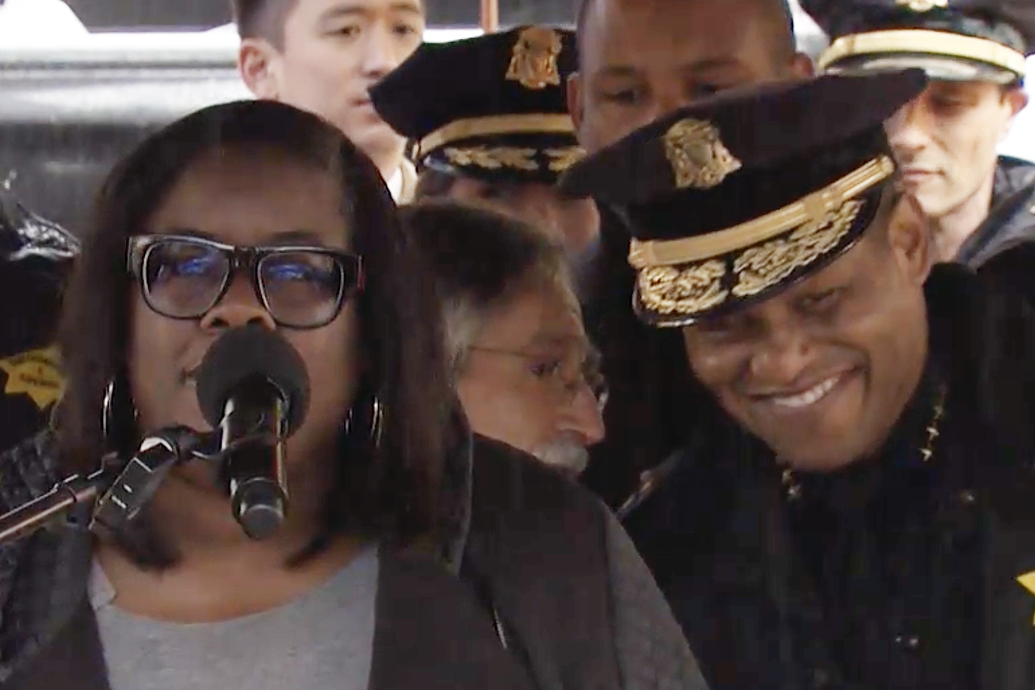 People at a press conference, with a woman at the microphone and a man behind her speaking with a police chief.