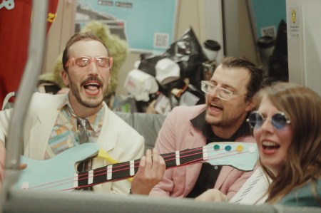 Three people laugh and sing, one playing a toy guitar, on a cluttered bus. They're joyful and casually dressed.