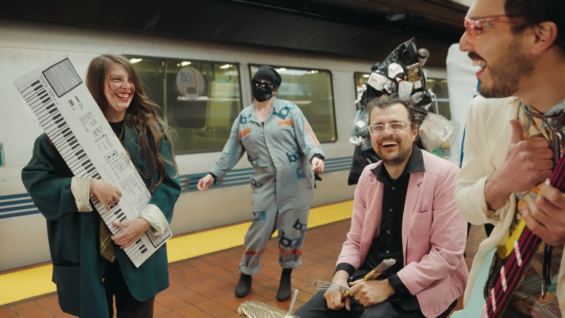 Band members carrying a papier-mache keyboard and electric bass smile at each other next to a drummer holding a whisk on a subway station platform, with a dancer standing nearby next to a train.