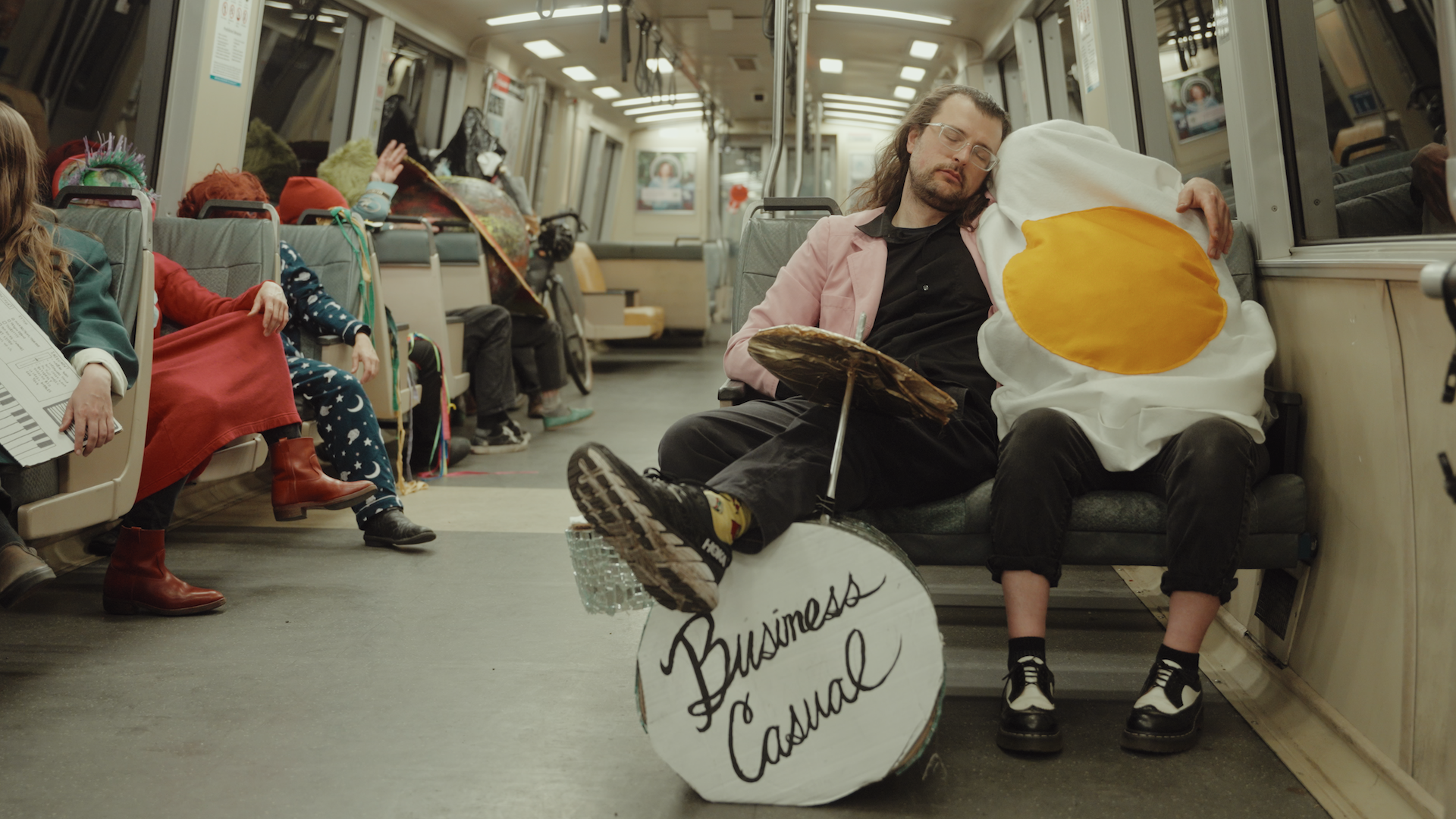 A man with long hair and glasses wearing a pink blazer over dark clothing rests his legs on a toy drum kit and drapes his left arm over a person wearing a fried egg costume. Both sit asleep on a subway train seat.