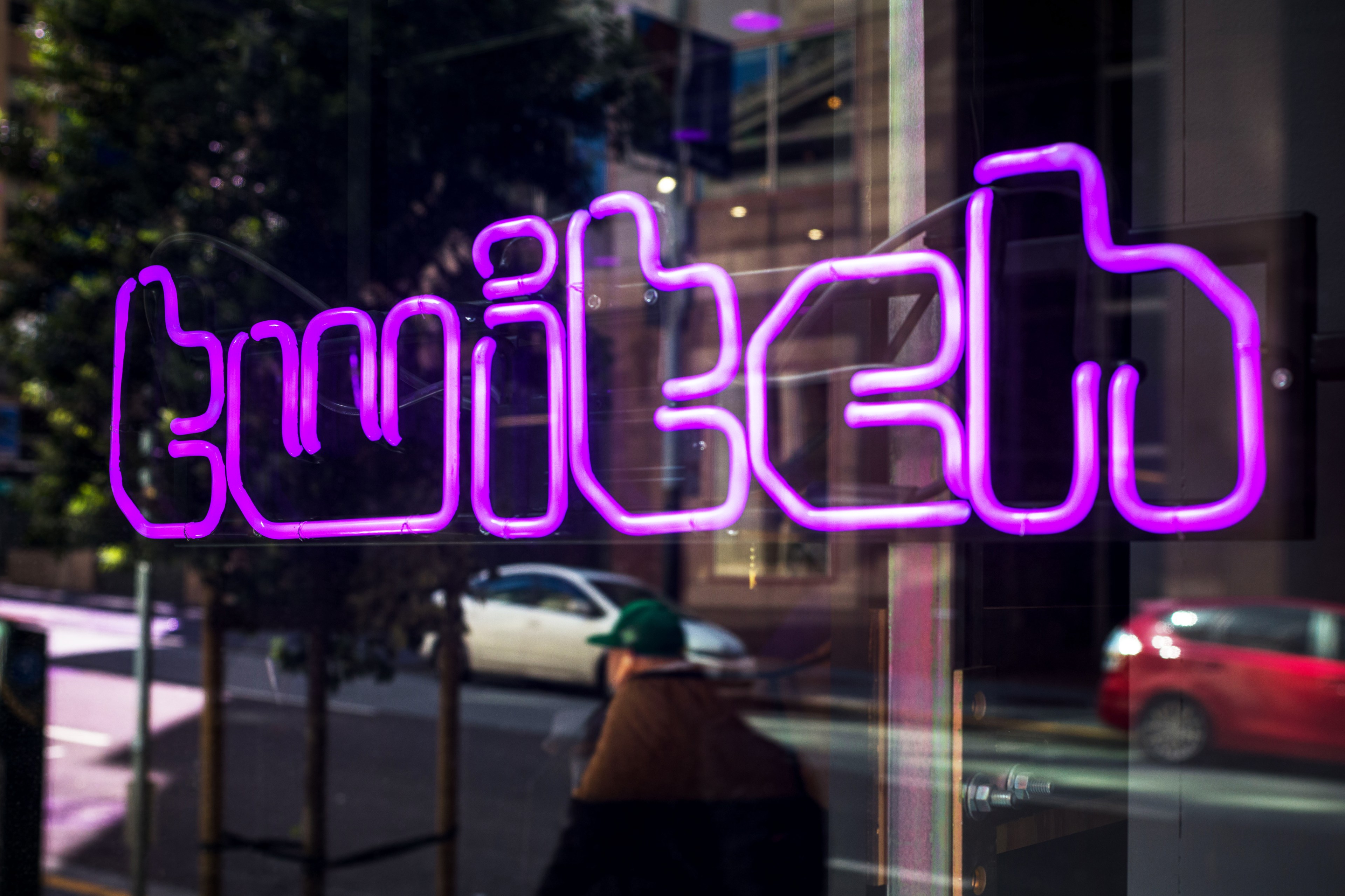 A neon sign with mirror-image text against a window, reflecting street life with moving cars and a pedestrian.