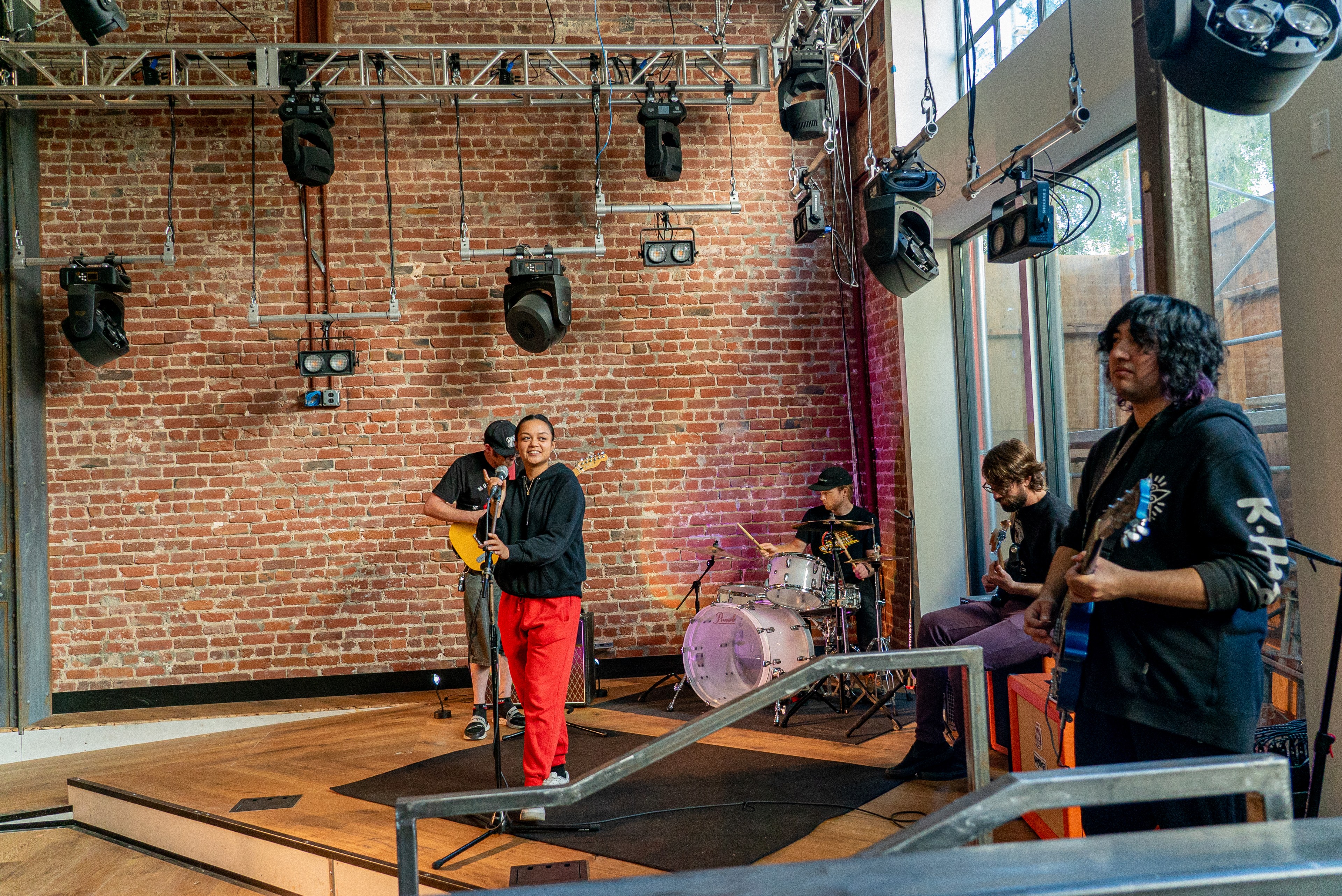 Musicians play guitars and drums in a performance space with wooden floors and a brick wall.