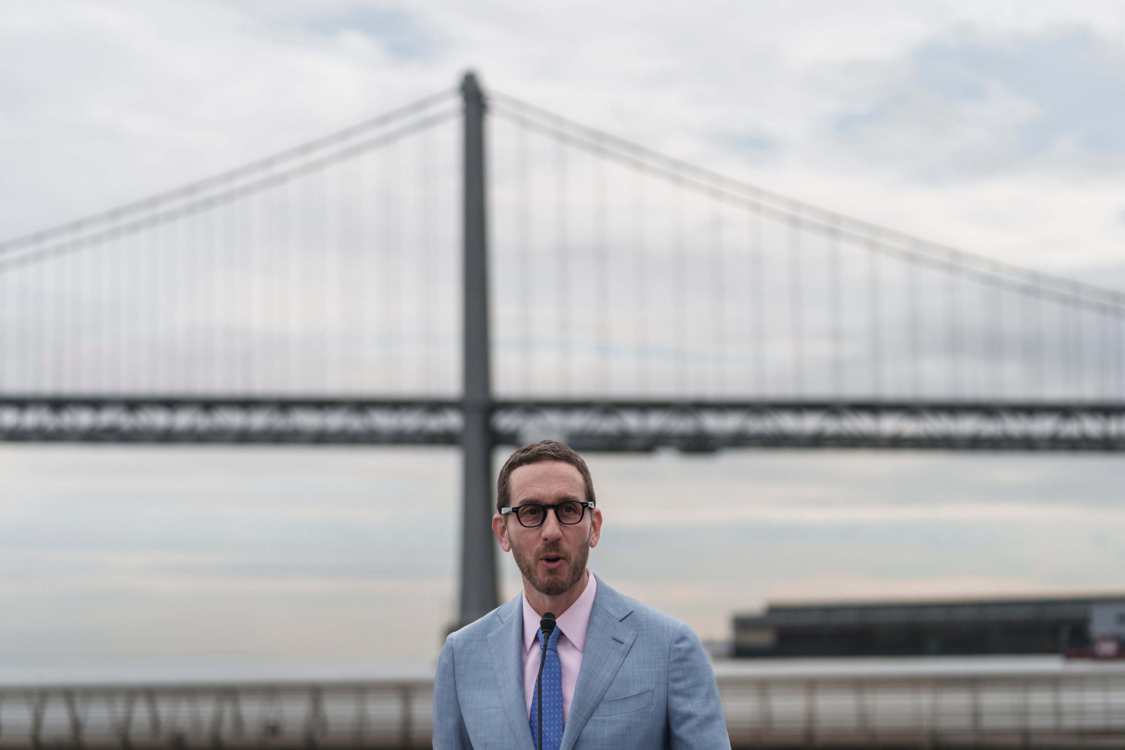 A man wearing glasses and a light blue suit stands in front of a large suspension bridge under a cloudy sky.