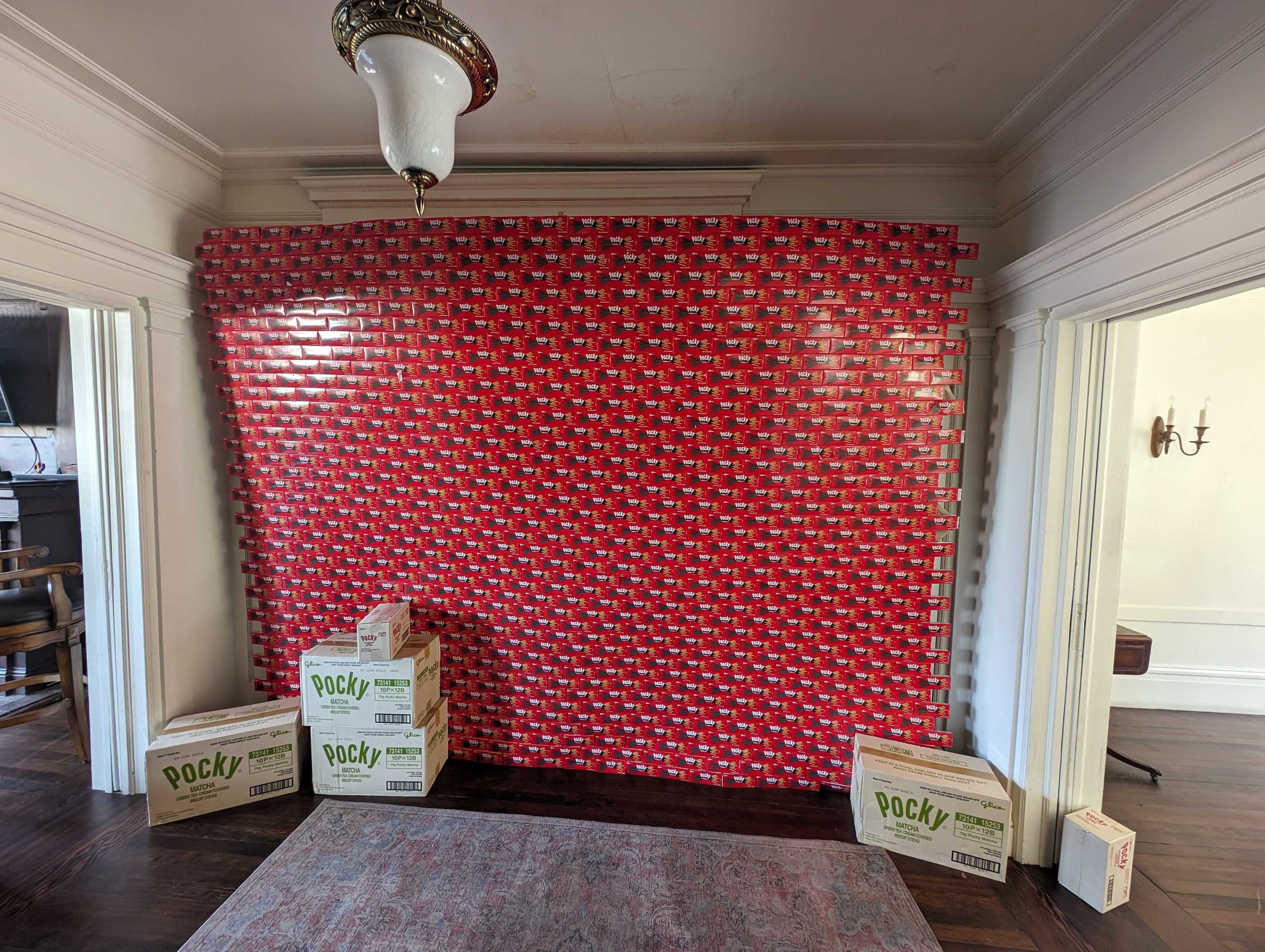 A display wall of empty chocolate-flavored Pocky cartons stands floor-to-ceiling in the lobby of a home, with several cardboard boxes of matcha-flavored Pocky at its base.