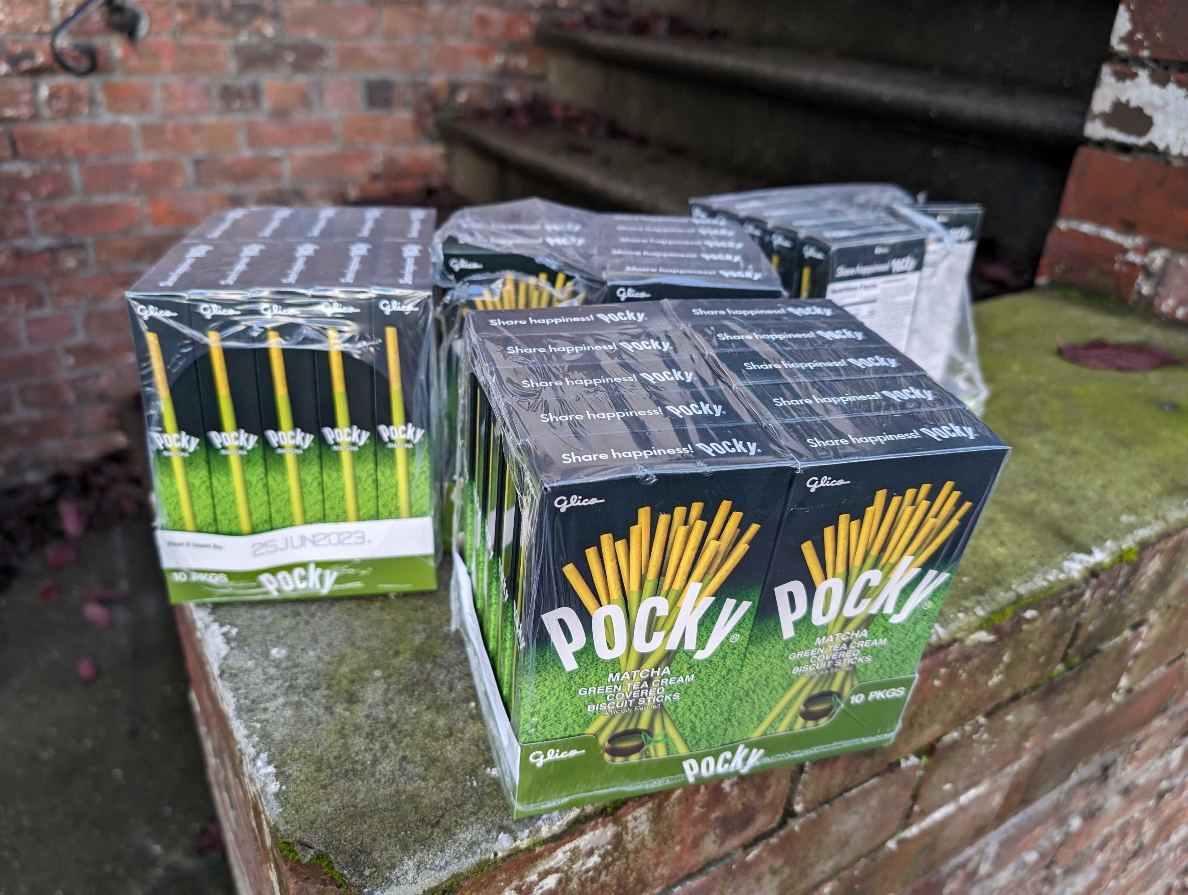 Four plastic-wrapped boxes of matcha green tea cream flavored Pocky biscuit sticks sit on a low brick stoop.