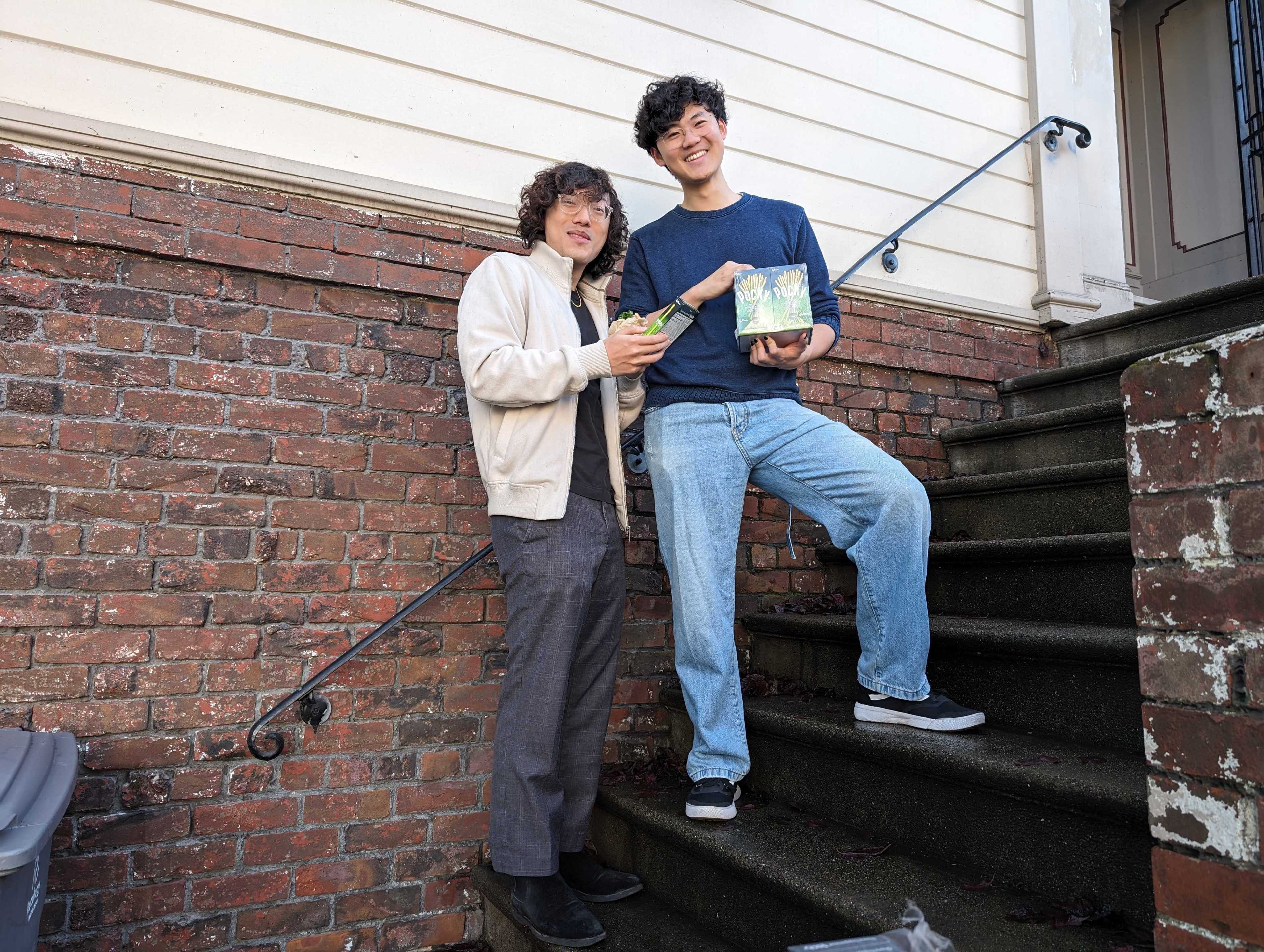 Two smling young men stand on a staircase outside a home in front of a brick wall and wooden siding. Both men hold boxes of Pocky biscuit treats.