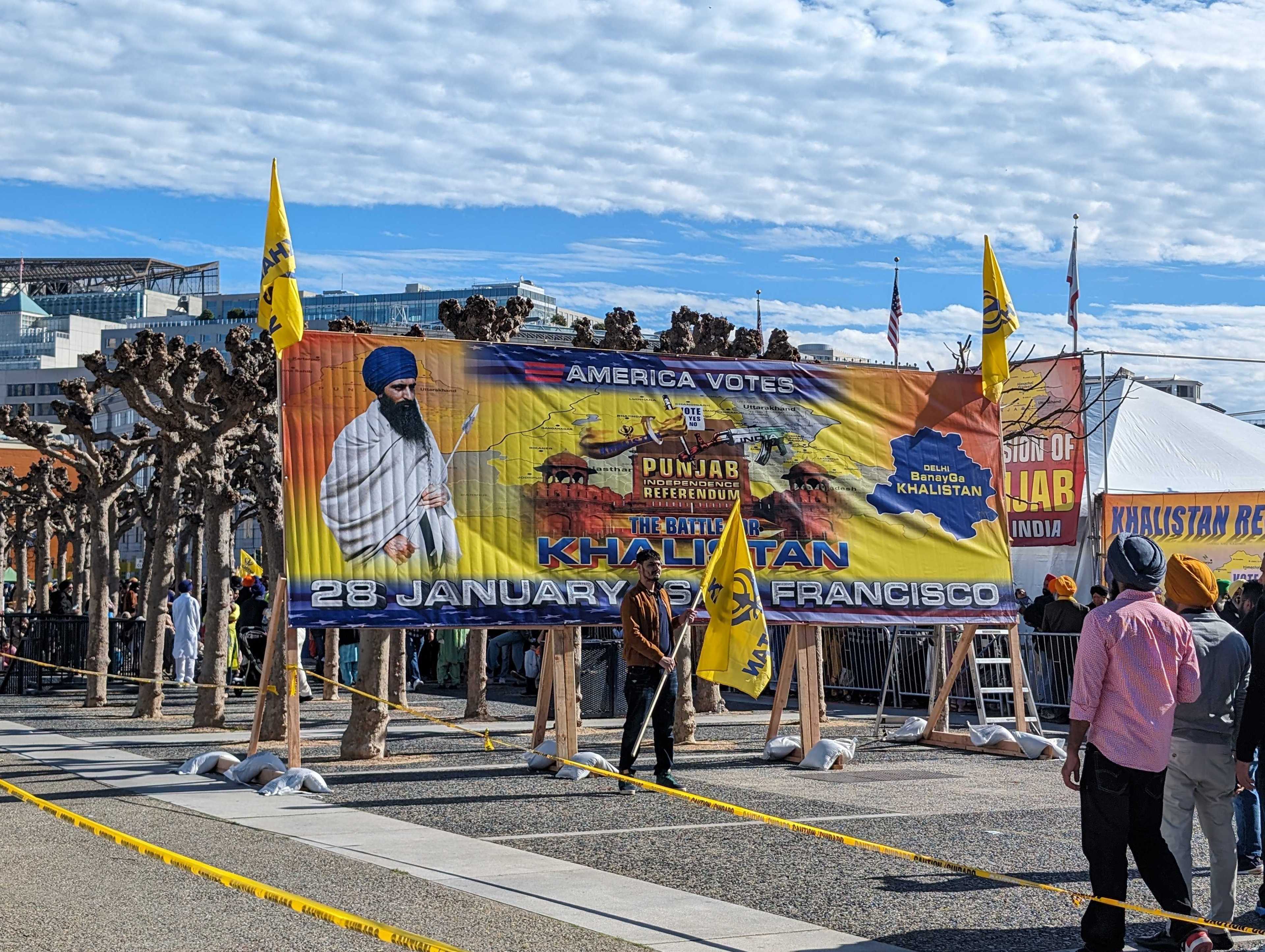 A man holds a yellow flag in front of a sign advertising a voting referendum at an outdoor public plaza.