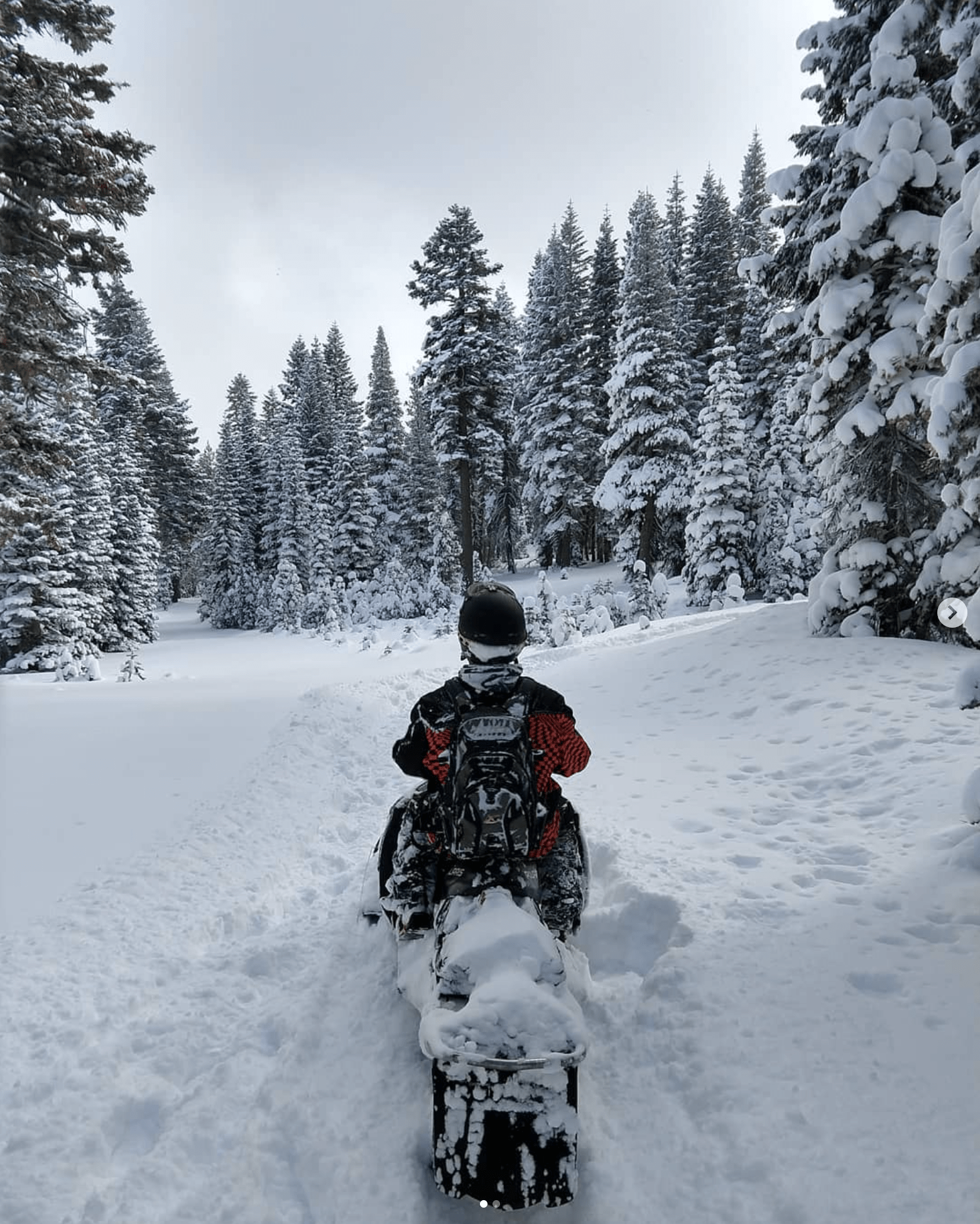 The back of a man on a snowmobile in a snowy, winter landscape with lots of powder on the ground and on his jacket.