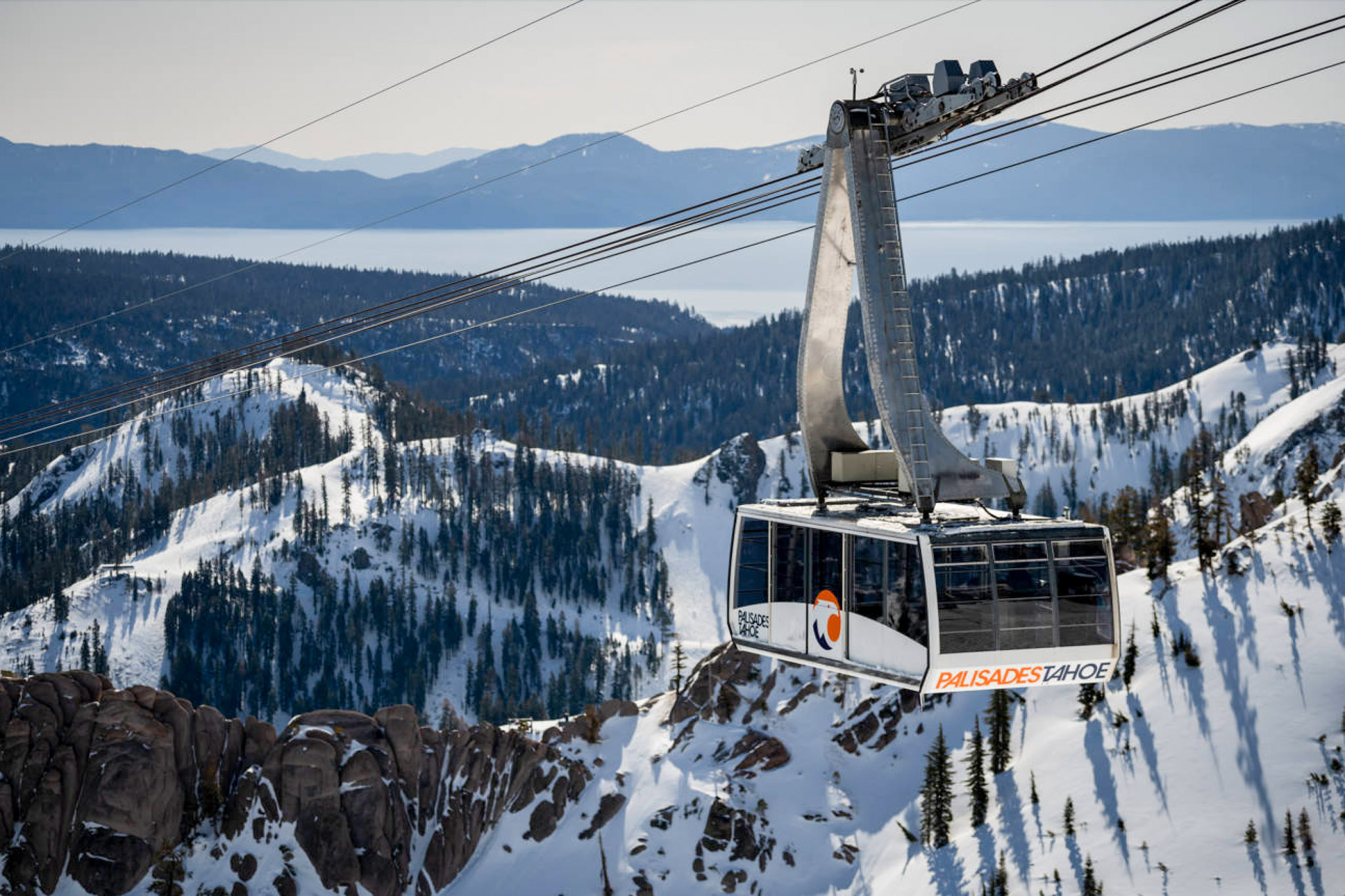 An aerial tram hangs from cables amidst a snowy winter scene with Lake Tahoe in the background.