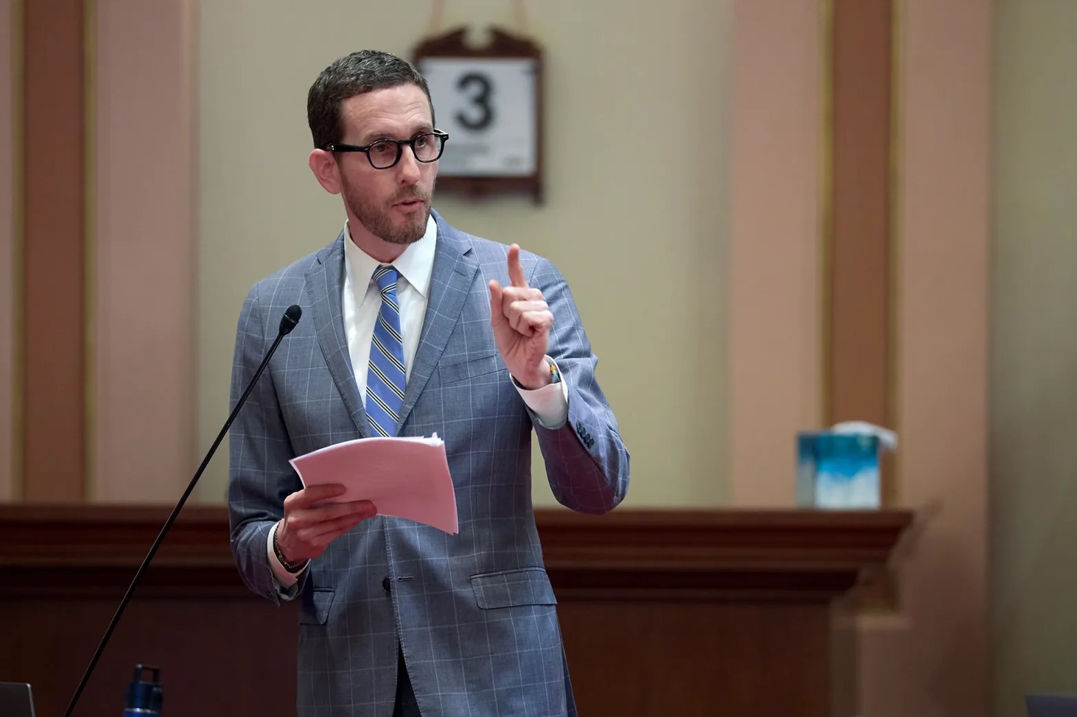 a man in a light blue suit holding a sheaf of papers points while speaking