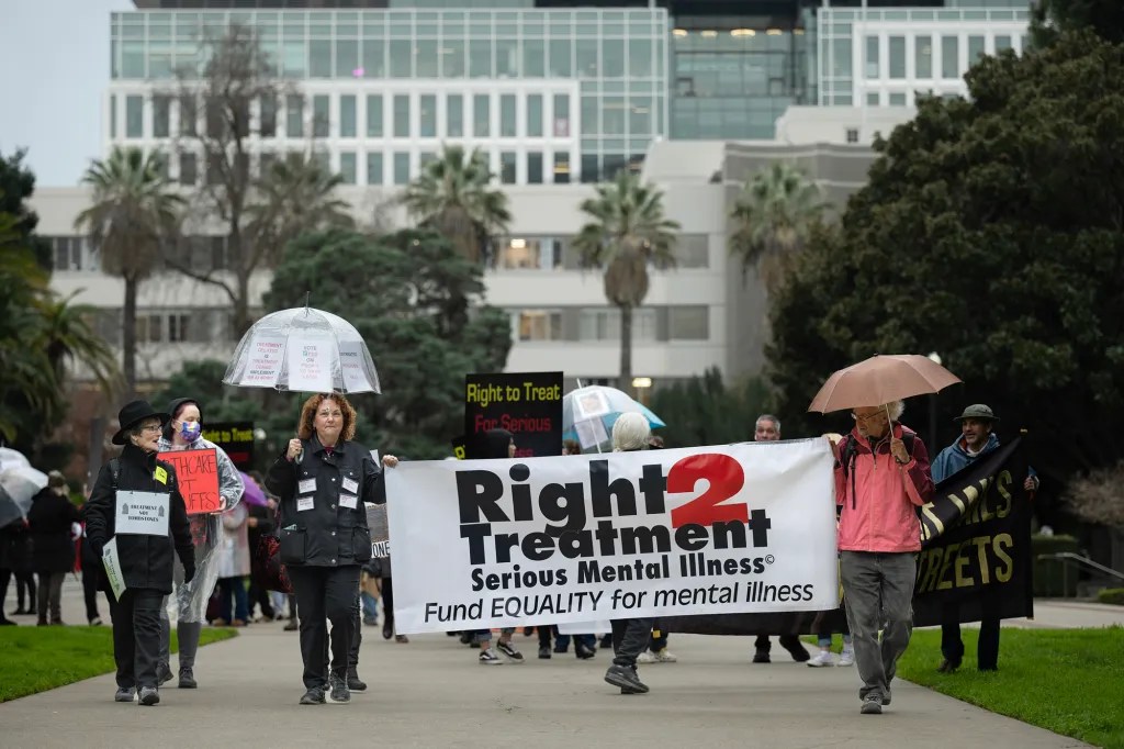 Man and woman walk carrying umbrellas and holding up banner that says Right 2 Treatment serious mental illness fund equality for mental illness