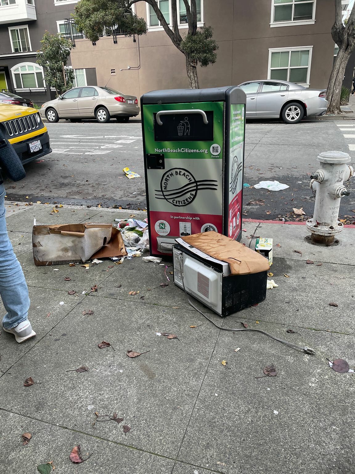 A microwave sits in front of a trash can on a public street.