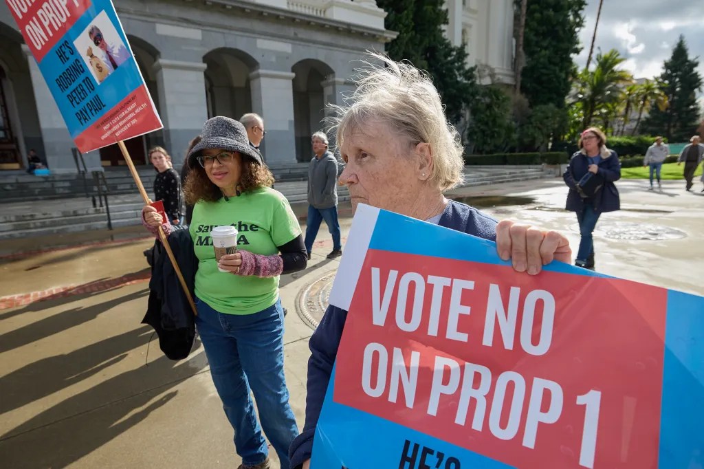 Two women hold signs with the words vote no on prop 1 as people walk behind them in front of gray building