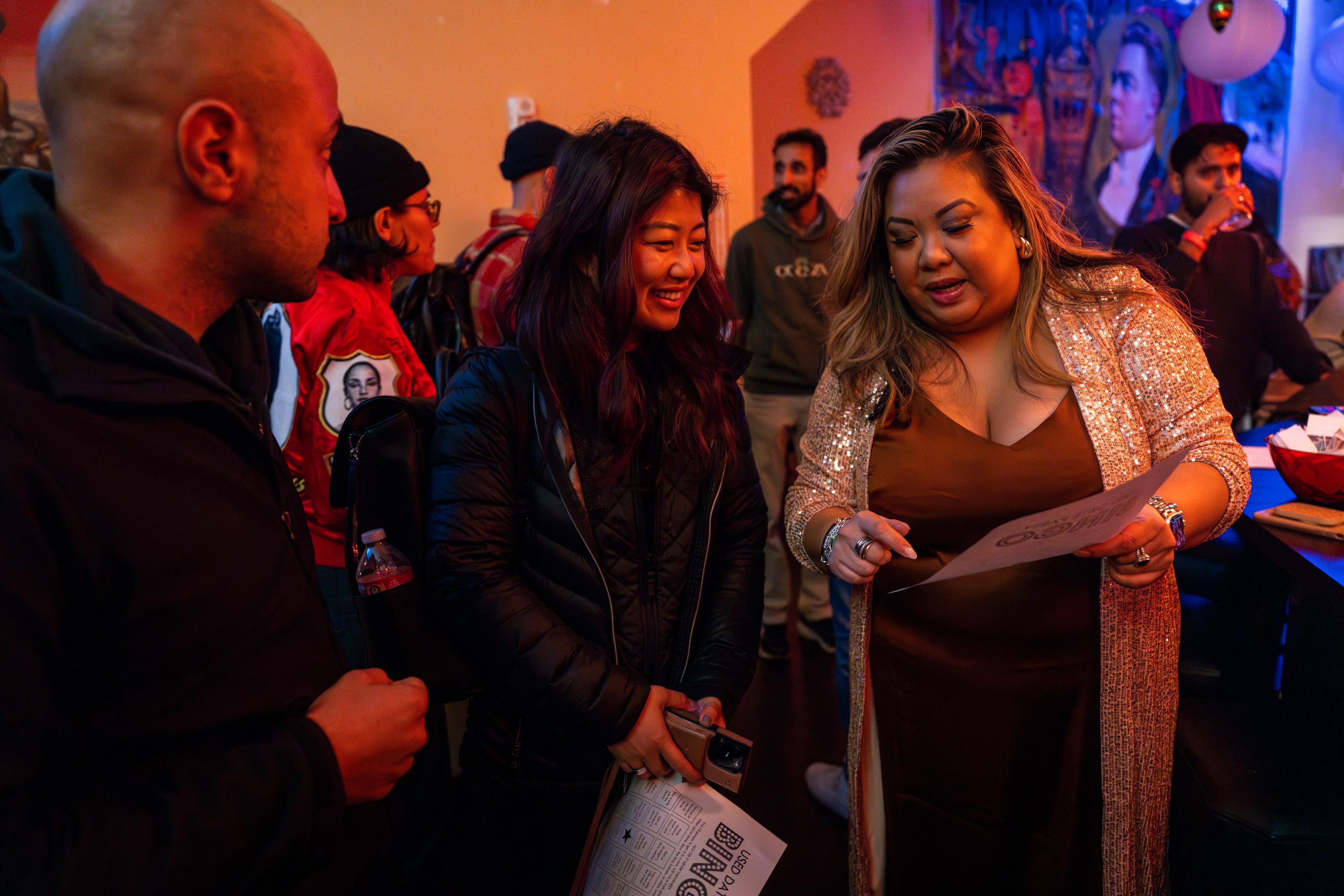 A woman shows a party attendee how to play &quot;Used Date Party&quot; bingo at a single mixers event in a low lit bar in San Francisco.