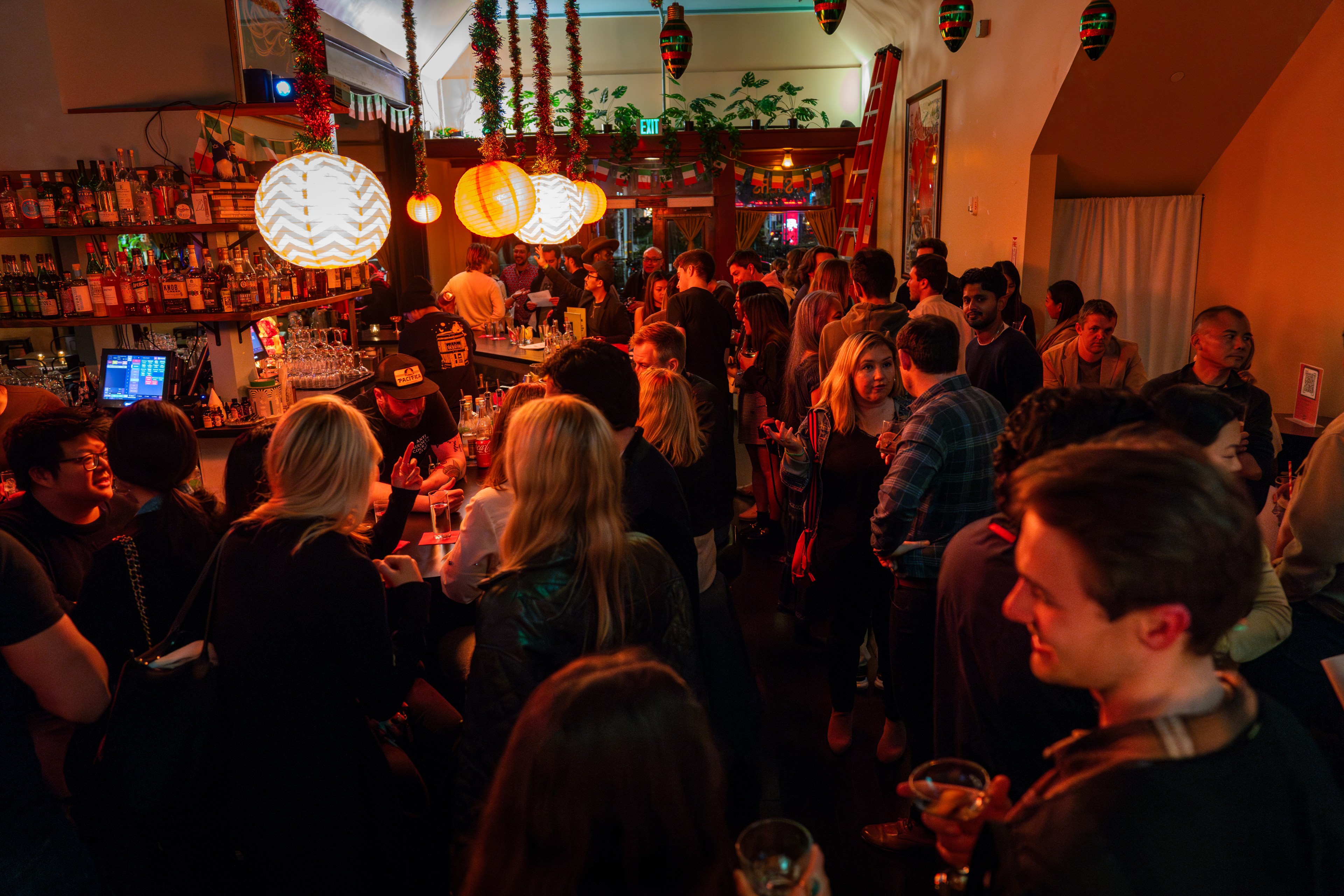 People gather in a crowded bar with low-hanging lights wrapped in tinsel and leftover Christmas ornaments hanging form the ceiling.