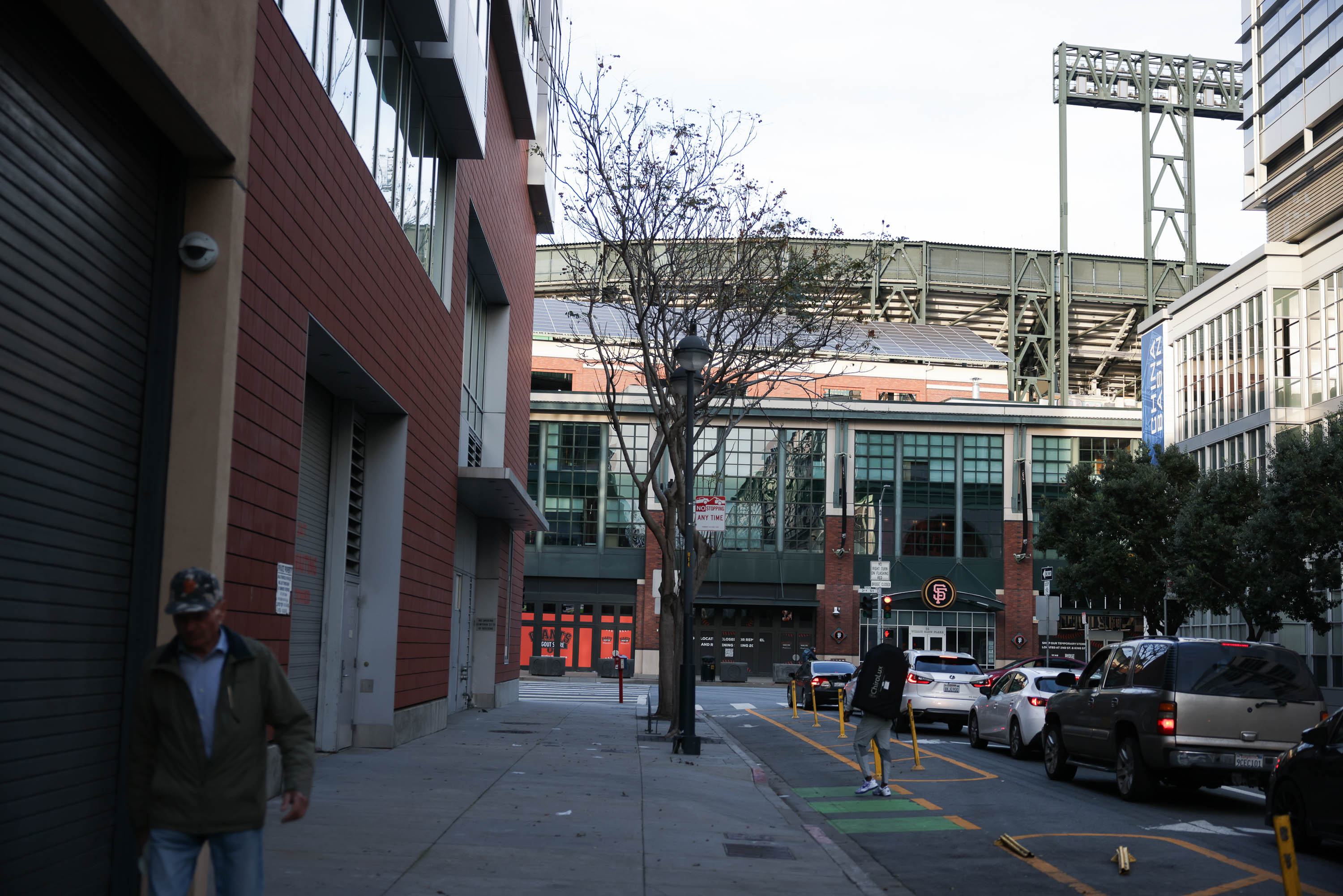 A street view with pedestrians, parked cars, and urban buildings leading to a stadium entrance.