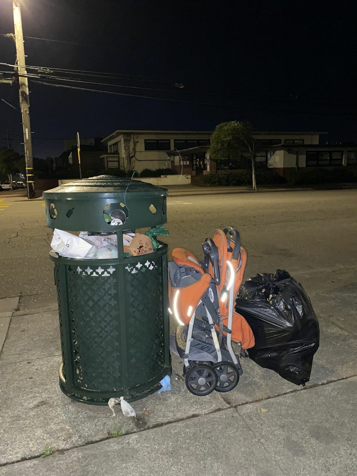 A stroller and a black bag of trash sit on the ground next to a public trash can.