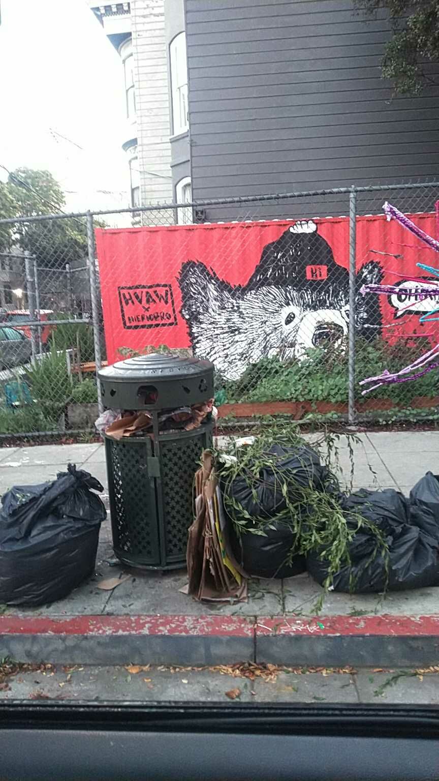 Black garbage bags with plants spilling out of them sit next to a trash can.