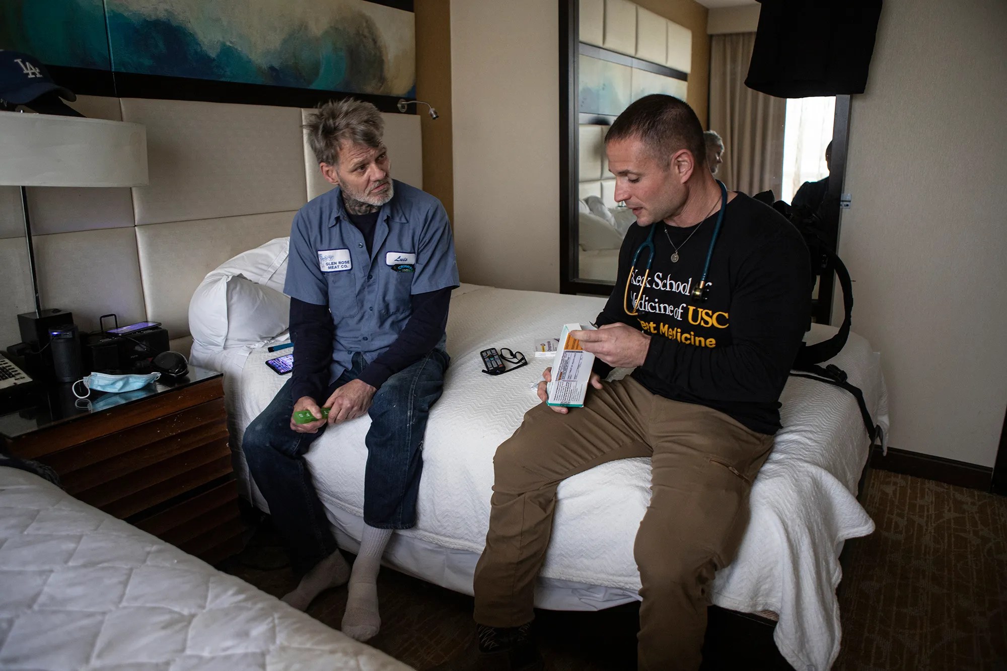 Two men sit on a hotel bed; one holds a medical device, the other looks on, with medical supplies scattered around.