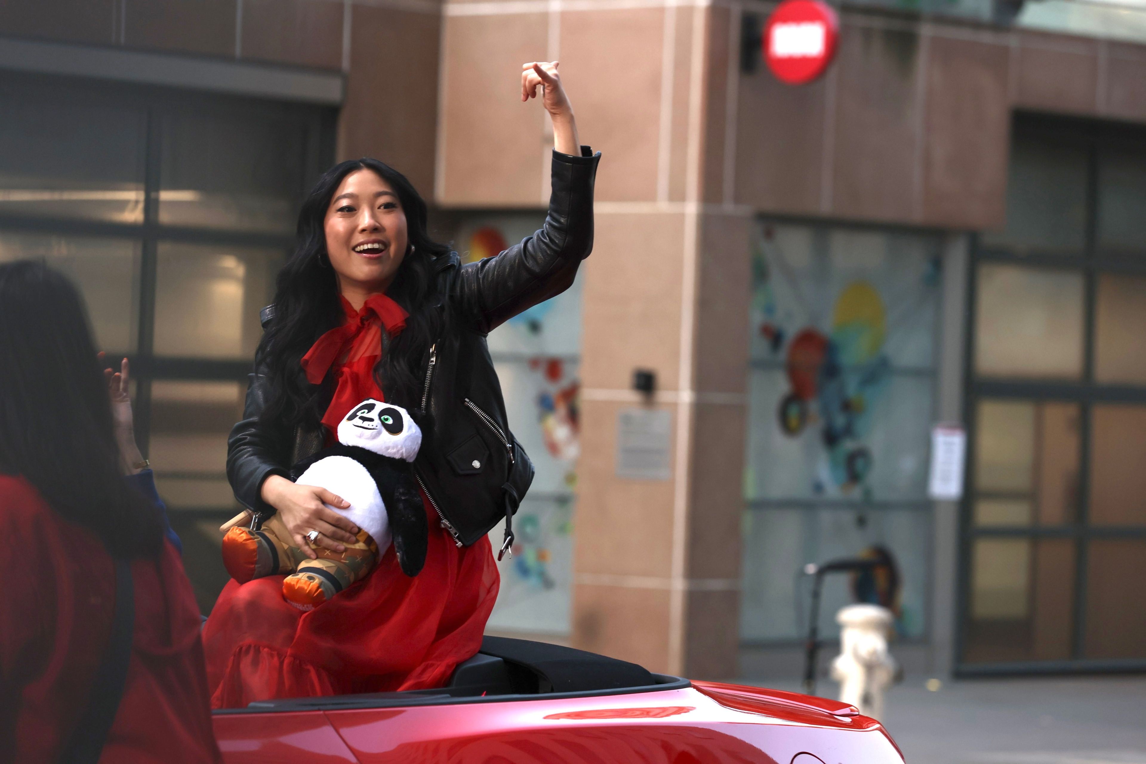 Woman holds panda in lap while riding atop a red car driving in a parade