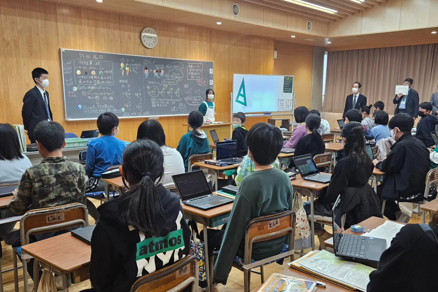 A classroom with Japanese students at desks, some with laptops, facing teachers near blackboards covered in writing.