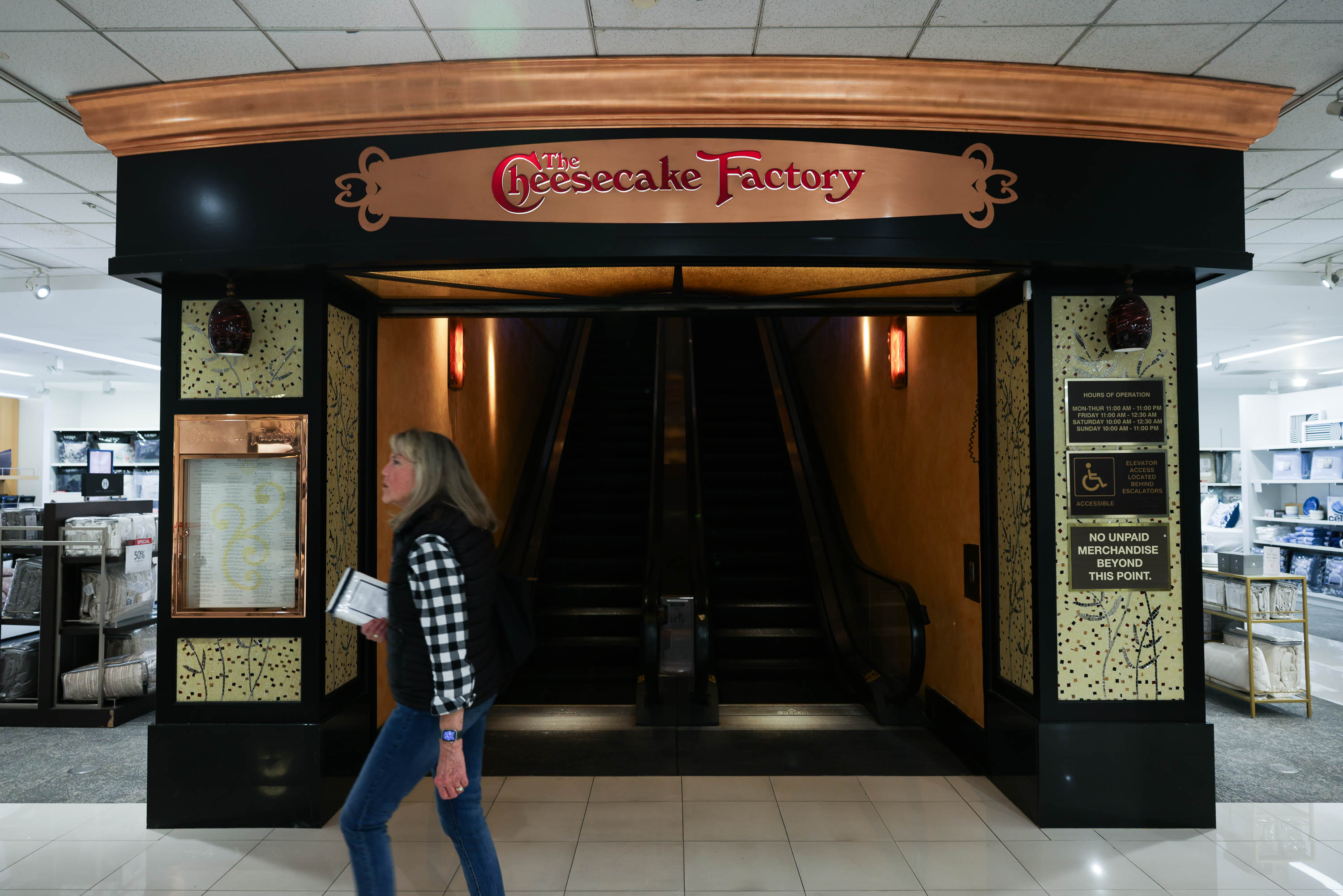 A person walks past the entrance to The Cheesecake Factory, with a menu displayed and an escalator inside.