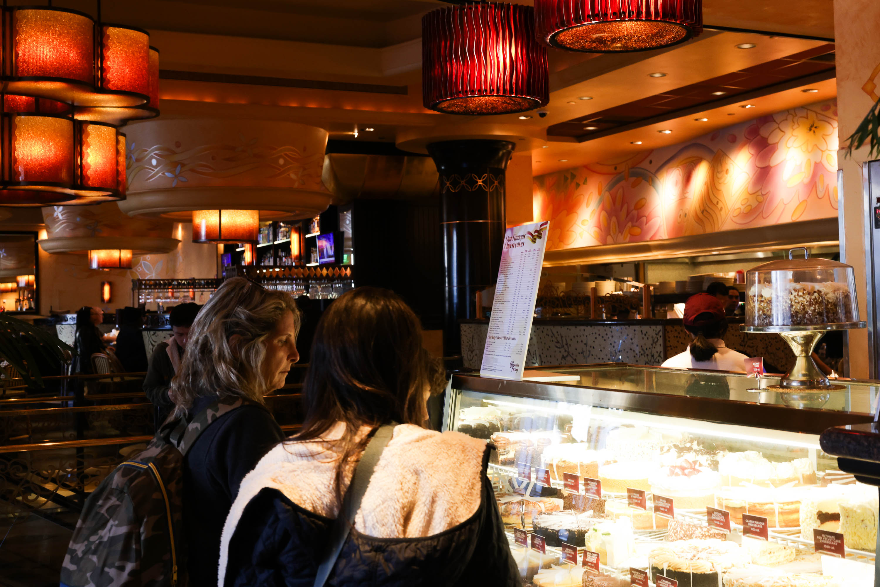 Two people gaze at a brightly-lit pastry display in a cozy bakery with warm ambient lighting.