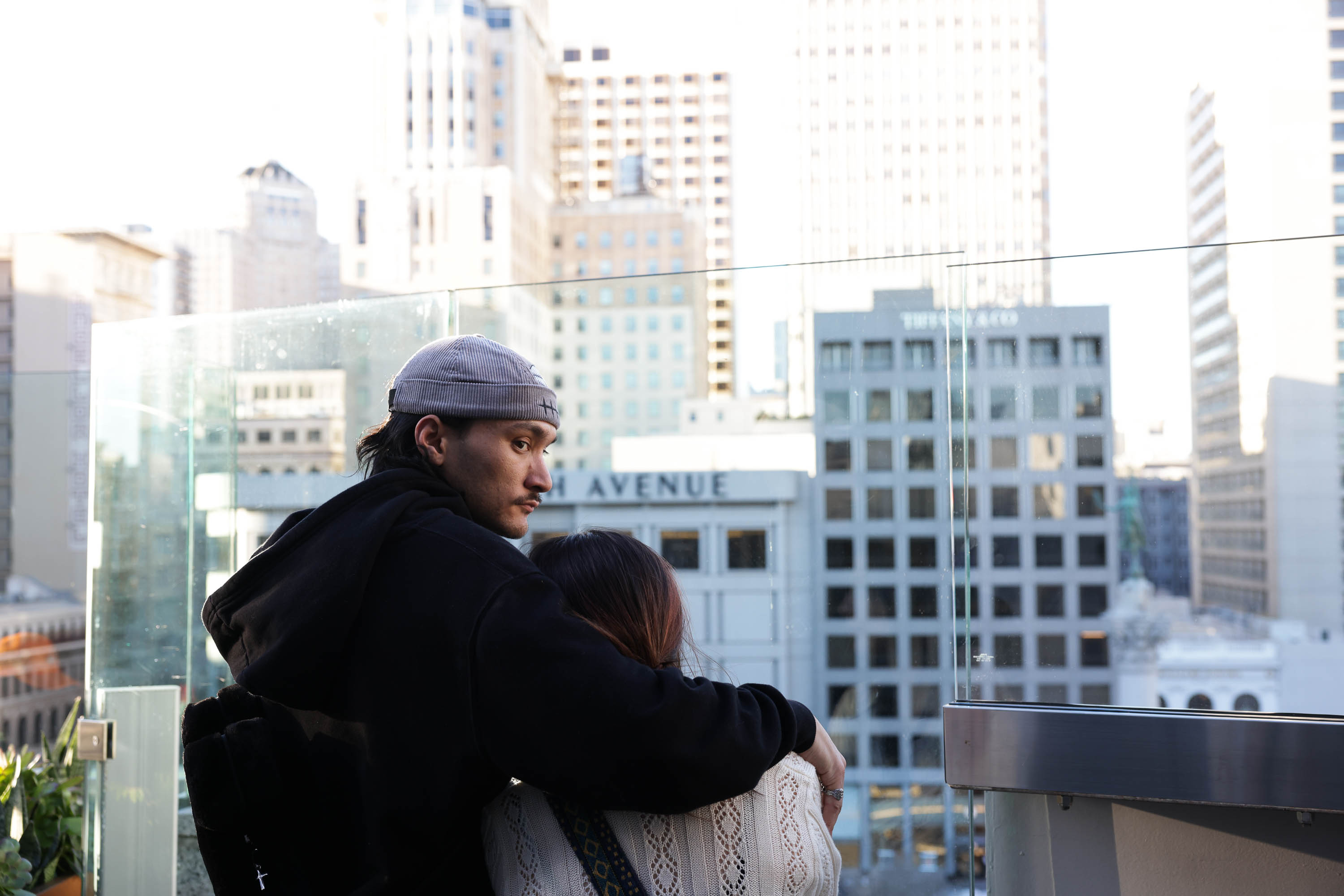 A man and woman are embracing on a balcony overlooking a cityscape.
