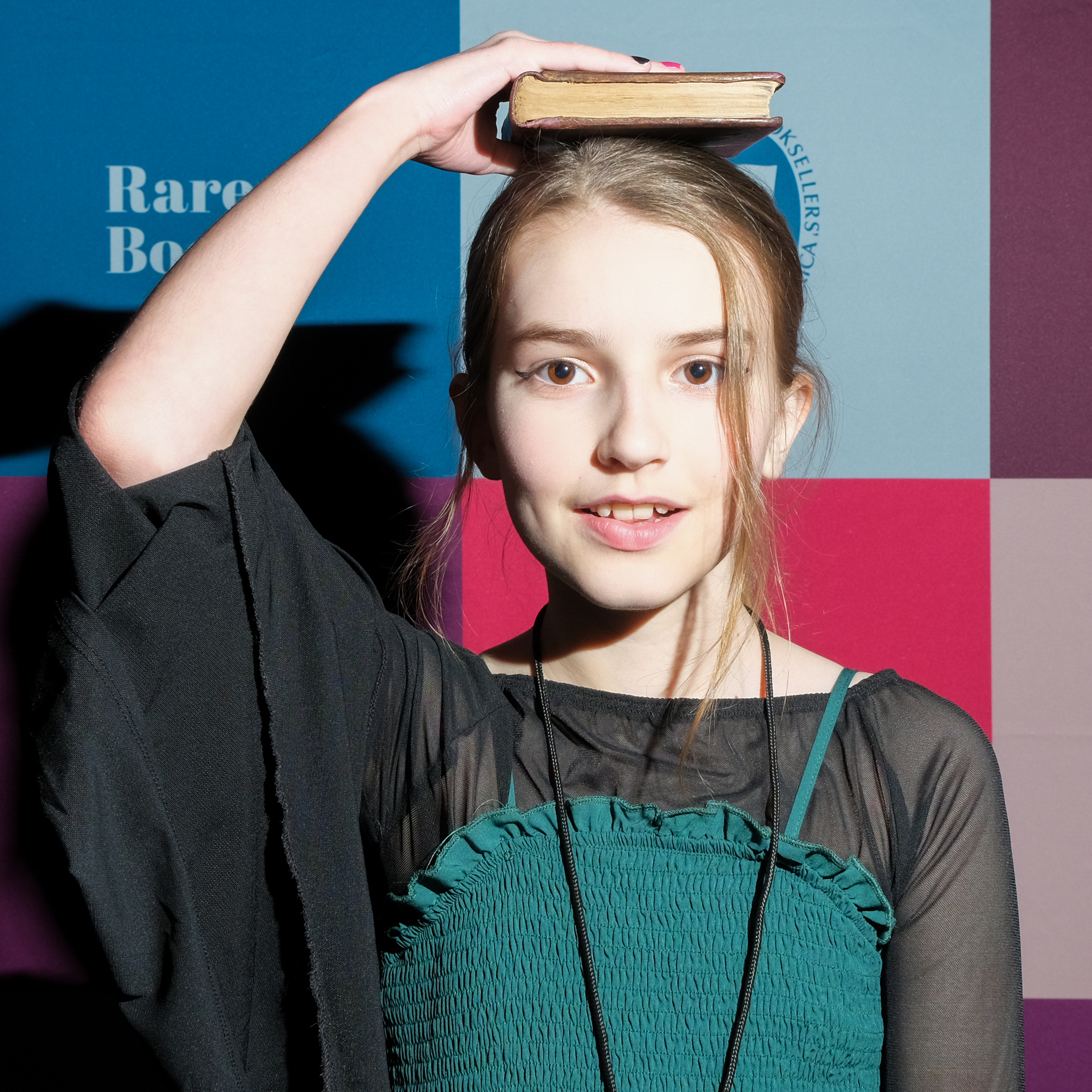 A girl balances a book on her head, wearing a black shirt, in front of a colorful backdrop.